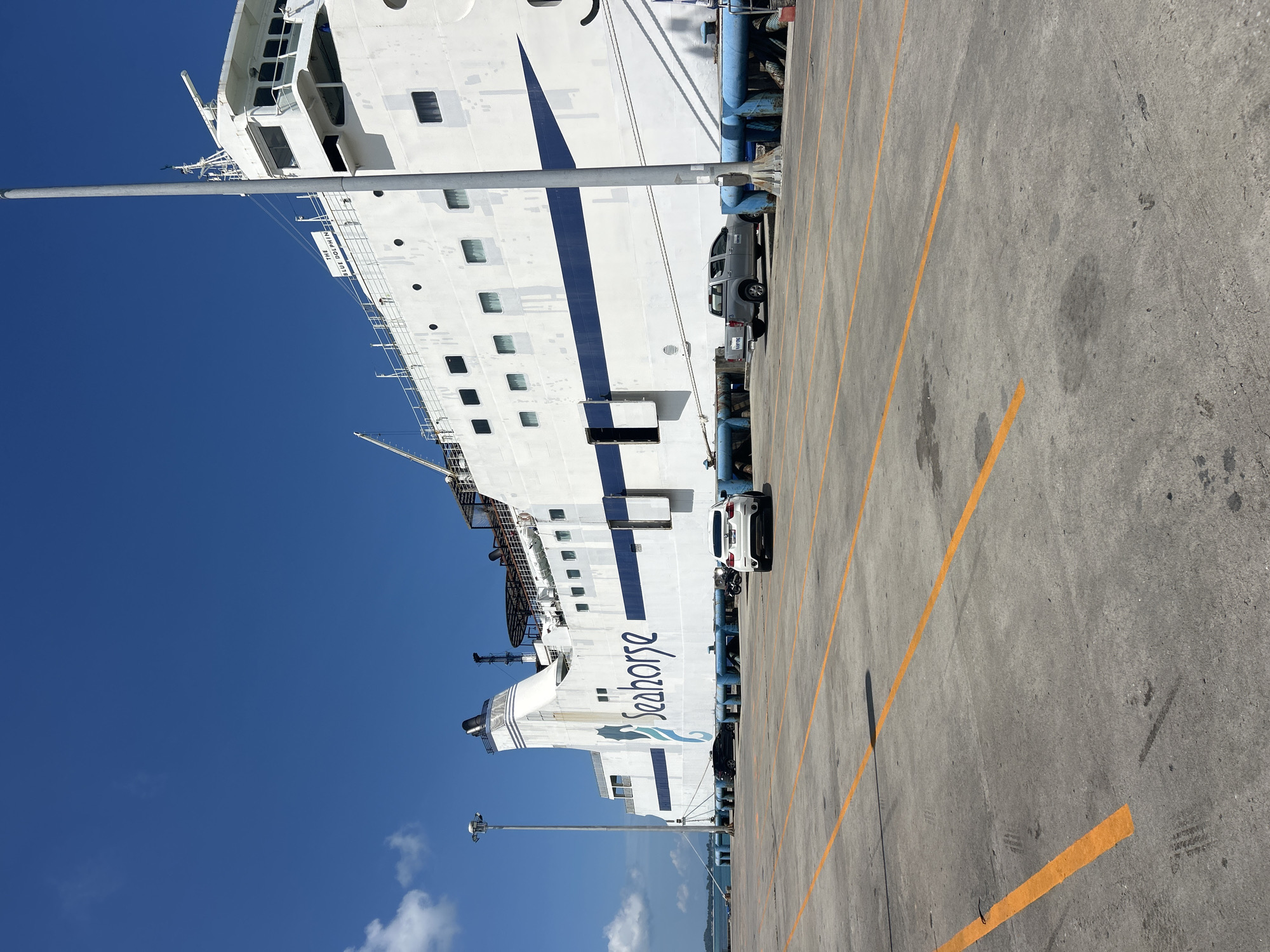A large white ferry, named "Seahorse", is docked at a pier in the city of Ko Samui District, Thailand. The ferry has blue trim and multiple windows. In front of the ferry, there is a concrete parking lot marked with yellow lines. Several vehicles are parked on the lot, including a white SUV and a silver pickup truck. The sky is clear and blue with a few scattered clouds. A tall light pole is visible on the left side of the image. The overall scene depicts a sunny day at a ferry terminal.