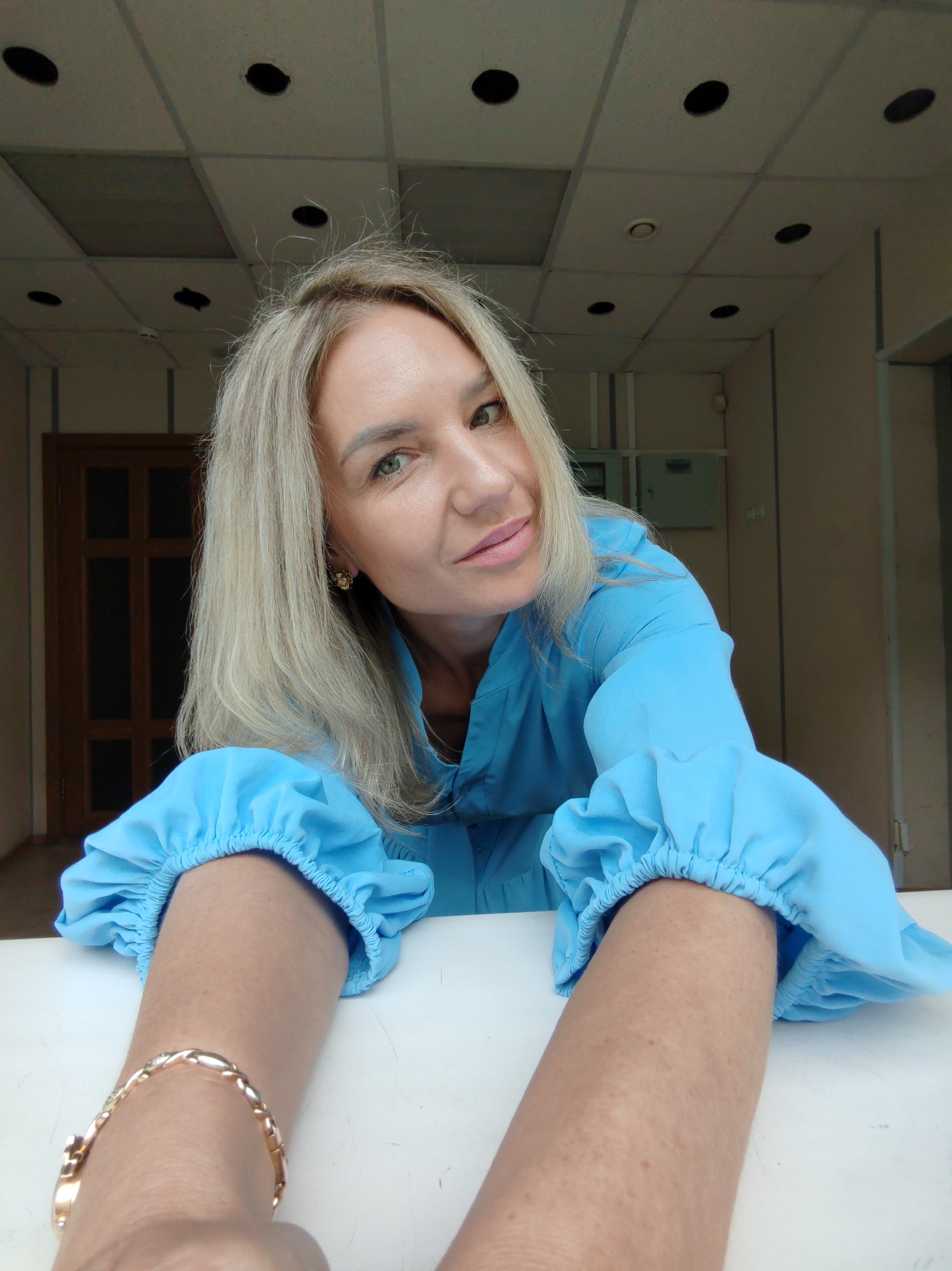 A woman with blonde hair, wearing a blue medical coat, poses for a selfie. Her hair is shoulder-length and slightly wavy. She has light skin and green eyes, and she is smiling slightly. The woman is leaning forward, her arms resting on a white surface. She is wearing a gold bracelet on her left wrist. The background is a room with a drop ceiling, light-colored walls, and a wooden door. The setting appears to be an office or medical facility. The image was taken indoors.
