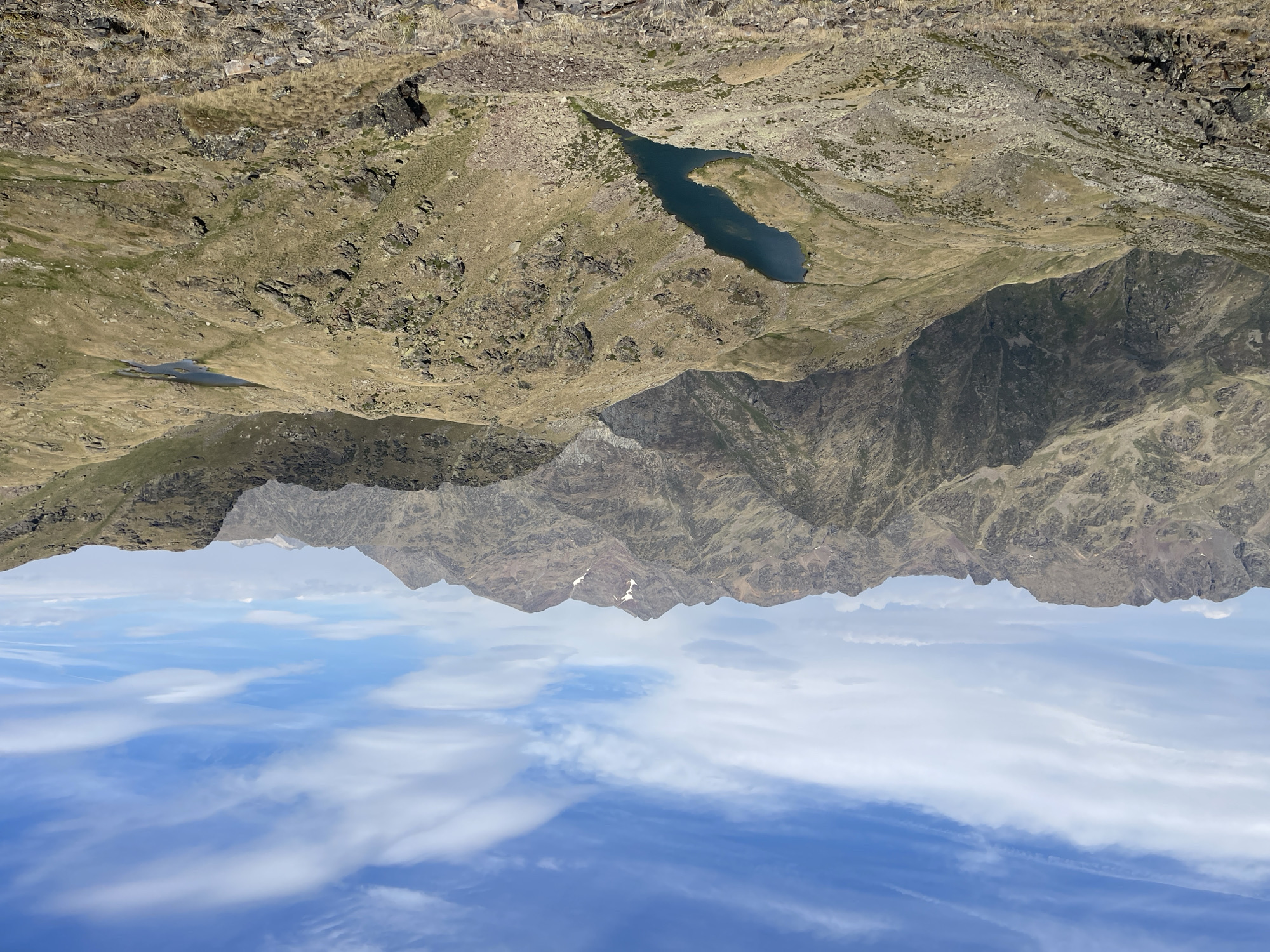 An eye-level, wide shot captures a vast mountain landscape under a bright blue sky with scattered white clouds. The foreground is dominated by rocky, dry terrain covered in sparse, yellow-brown grass, with several large rocks and boulders. In the mid-ground, two alpine lakes are visible: a larger, elongated dark blue lake in the center-left, nestled in a valley, and a smaller, rounder dark blue lake further to the right, situated on a flatter, grassy area. Beyond the lakes, a series of rugged mountains rise, with varying shades of brown and gray rock, some slopes appearing steep and barren, others covered in patches of green and yellow vegetation. The distant mountain range is more muted in color, shrouded in a slight atmospheric haze, with a prominent peak in the center possibly bearing a small patch of snow. The overall impression is of a wild, remote, and serene natural environment.