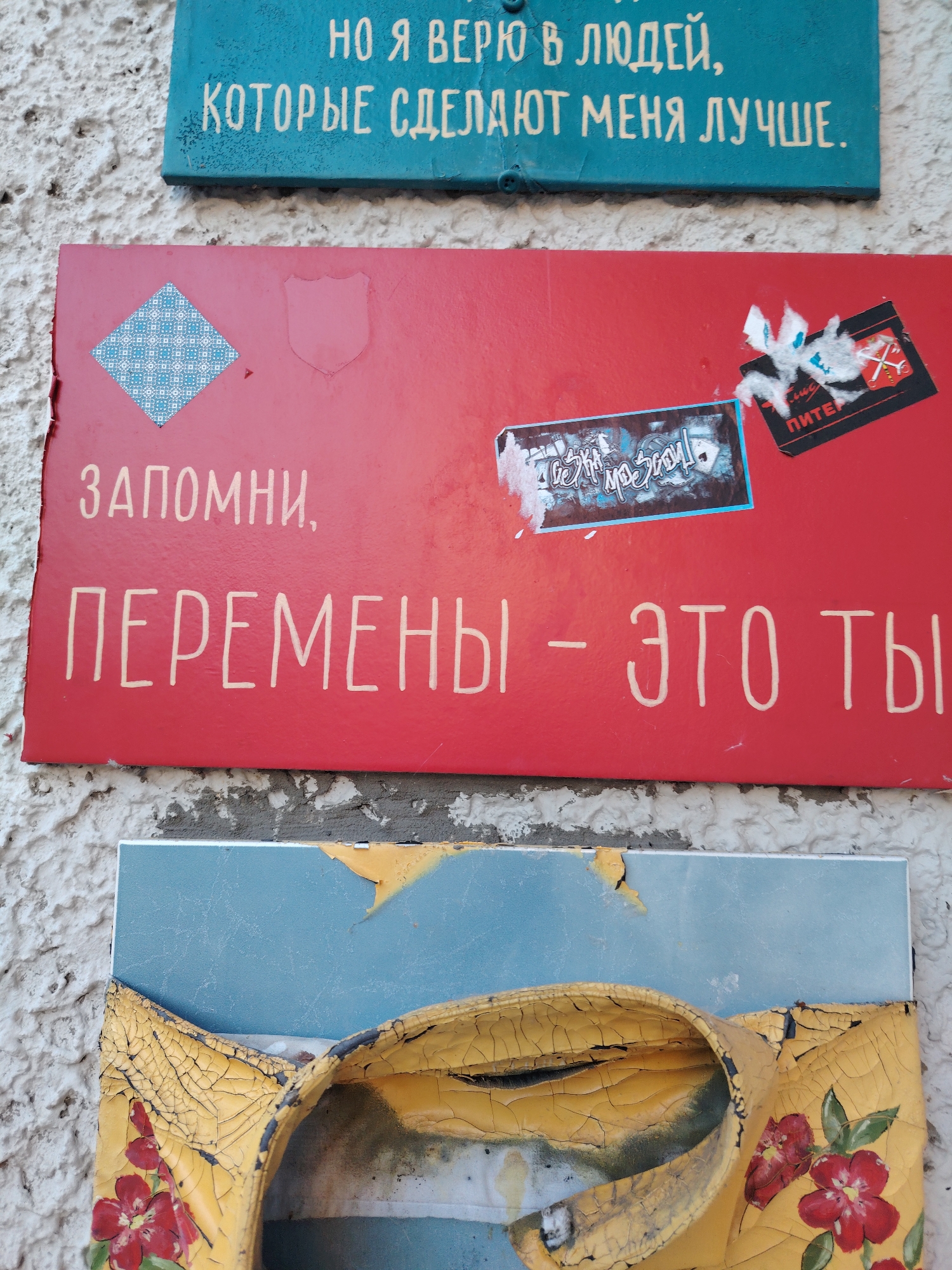 The image displays three weathered plaques attached to a textured, light-colored wall. The top plaque, teal in color, bears white Cyrillic text that reads, "НО Я ВЕРЮ В ЛЮДЕЙ, КОТОРЫЕ СДЕЛАЮТ МЕНЯ ЛУЧШЕ." (But I believe in people who will make me better.) The middle plaque is red and features white Cyrillic text, "ЗАПОМНИ, ПЕРЕМЕНЫ - ЭТО ТЫ." (Remember, changes are you.) A small blue and white patterned diamond shape is adhered to the upper left of this plaque, and to the right, a black sticker with a red and white emblem and the text "ПИТЕР" is partially torn. The bottom plaque is blue and yellow, with the yellow section peeling and cracked, revealing a floral pattern with red flowers. The plaque appears to be a decorative element, possibly a sign or artwork, in Konakovo, Russia. The scene is illuminated by natural light, casting subtle shadows. No people are visible in the image.