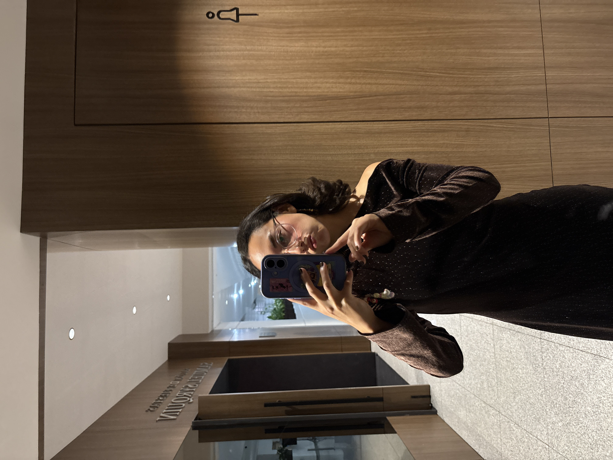 A young woman is captured taking a mirror selfie in a well-lit indoor environment. She has dark hair, wears glasses, and is dressed in a dark, shimmering long-sleeved top or dress. She holds a blue/purple smartphone with a case in both hands, angled towards a reflective surface. Her expression is neutral, focused on the phone.
The immediate background behind her reflection features wooden paneling with a horizontal grain. Prominently displayed on this paneling is a minimalist pictogram indicating a restroom, depicted as a stylized figure with a head and bell-shaped body. The floor in the reflection appears to be tiled with light-colored, possibly gray, square tiles.
Beyond the woman and the restroom sign, the reflection reveals a modern interior. White walls are visible, illuminated by several uniformly spaced circular recessed lights. Below these walls, a wooden counter or ledge features raised metallic lettering. When viewed correctly (un-mirrored), this text spells "HUIXQUILUCAN", which corresponds to the provided location context of Huixquilucan de Degollado, Mexico. The scene suggests a contemporary building, possibly a hotel, office, or event venue. No specific time of day or weather can be determined from the indoor setting.