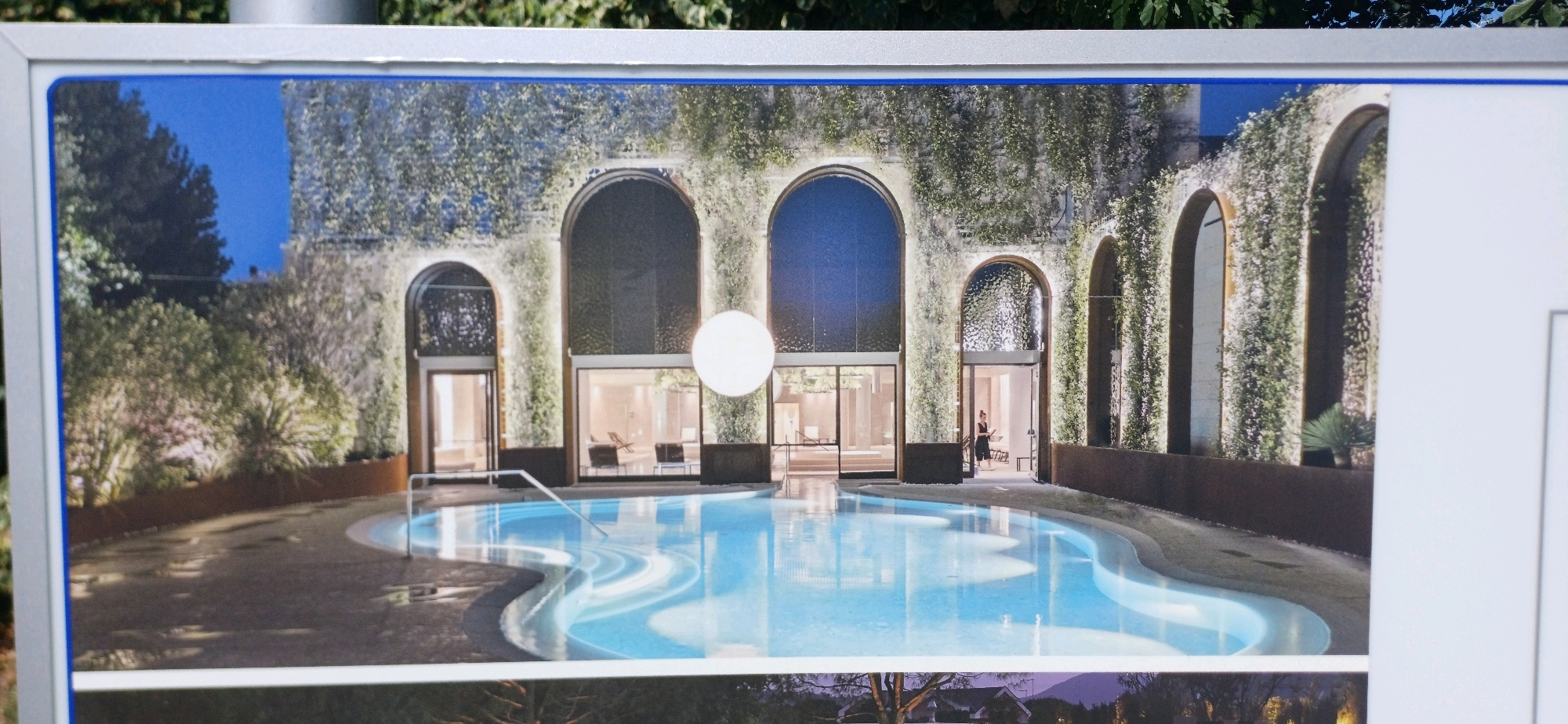 The image depicts an architectural rendering of a modern outdoor swimming pool area adjacent to a building with arched entrances and windows, heavily covered in lush green ivy. The pool itself is irregularly shaped, with a bright blue, illuminated water surface and a stepped entry. In the background, through the arched openings and windows, interior spaces with minimalist furniture are visible, including lounge chairs. A circular, illuminated light fixture hangs prominently in the air in front of one of the arched sections. A person in dark clothing is visible standing within one of the interior spaces near an arched doorway. The overall scene suggests a tranquil, luxurious resort or spa setting. The provided location context states this is from Abano Terme, Italy, a town known for its thermal spas, which aligns with the visual representation of a serene and possibly therapeutic water feature.