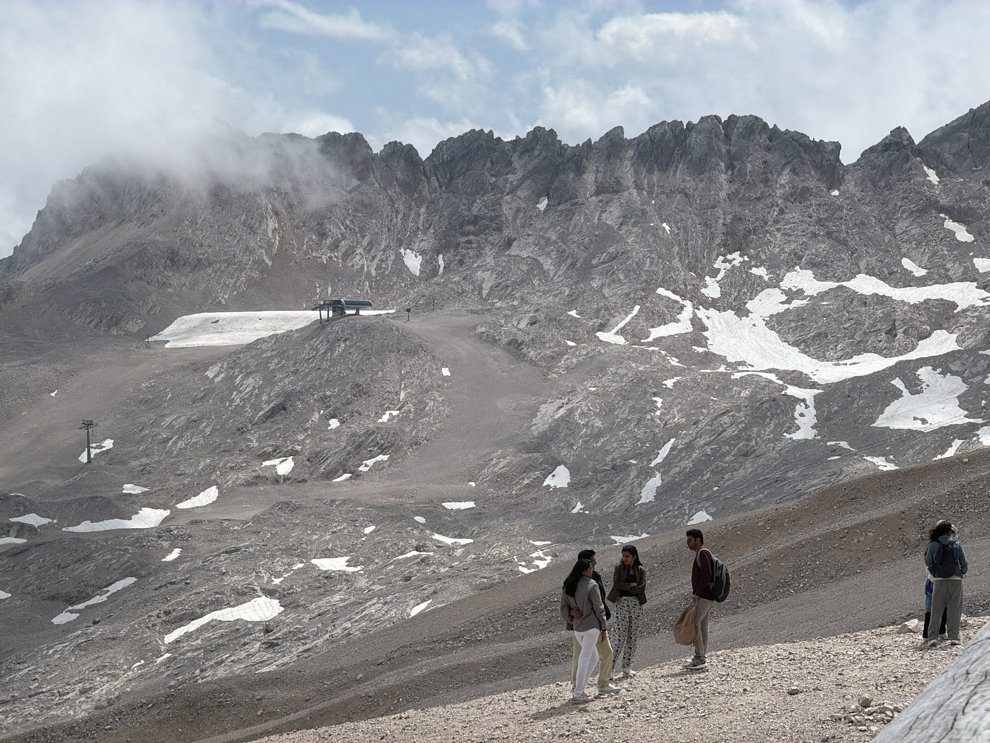 This is a photograph of the Zugspitze, the highest peak in Germany, located near Garmisch-Partenkirchen. The scene depicts a mountainous landscape with a cable car station visible in the distance, perched on a rocky outcrop. Patches of snow are scattered across the grey, rocky terrain. In the foreground, a group of four people are standing and looking towards the mountains. Two individuals are facing each other, engaged in conversation, while the other two stand slightly apart, observing the scenery. The sky above is partly cloudy with a mix of blue sky and white clouds. The overall impression is of a scenic mountain vista with tourists enjoying the view.