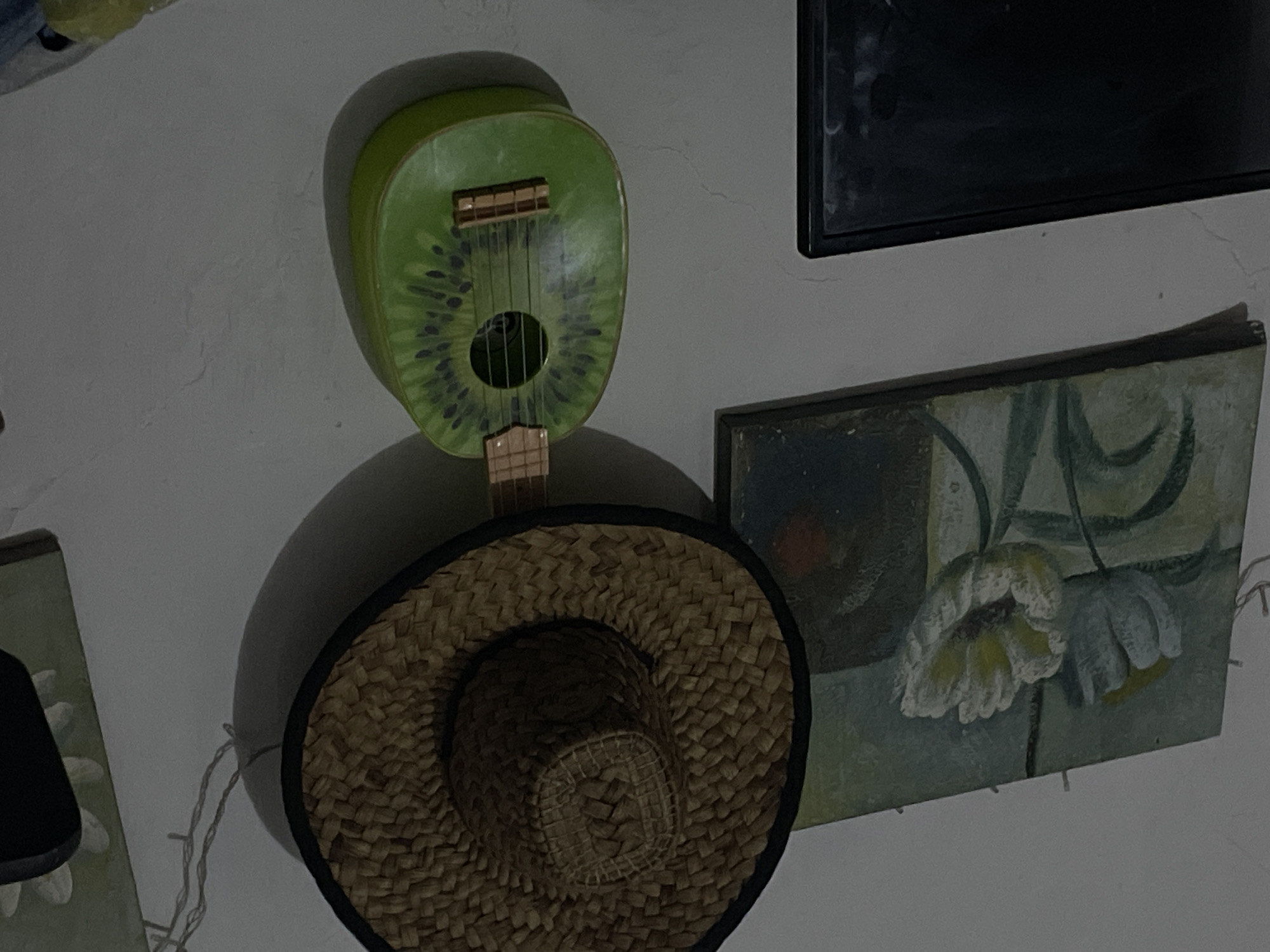 The image shows a collection of objects displayed on a textured, off-white wall, likely indoors. Dominating the center is a novelty ukulele shaped and decorated to resemble a kiwi fruit, complete with seeds. Below the ukulele, a woven straw hat with a black rim is suspended, casting a shadow on the wall. To the right of the ukulele and hat is a framed painting. The painting depicts a stylized floral subject, with white and yellow flowers rendered in a somewhat abstract manner against a muted green and blue background. In the upper right corner, a dark, rectangular object, possibly a television screen or another framed item, is partially visible. A string of small, decorative lights drapes across the wall, originating from behind the straw hat and extending downwards. The lighting in the scene is dim, suggesting it could be evening or a poorly lit room. There is no visible text in the image.