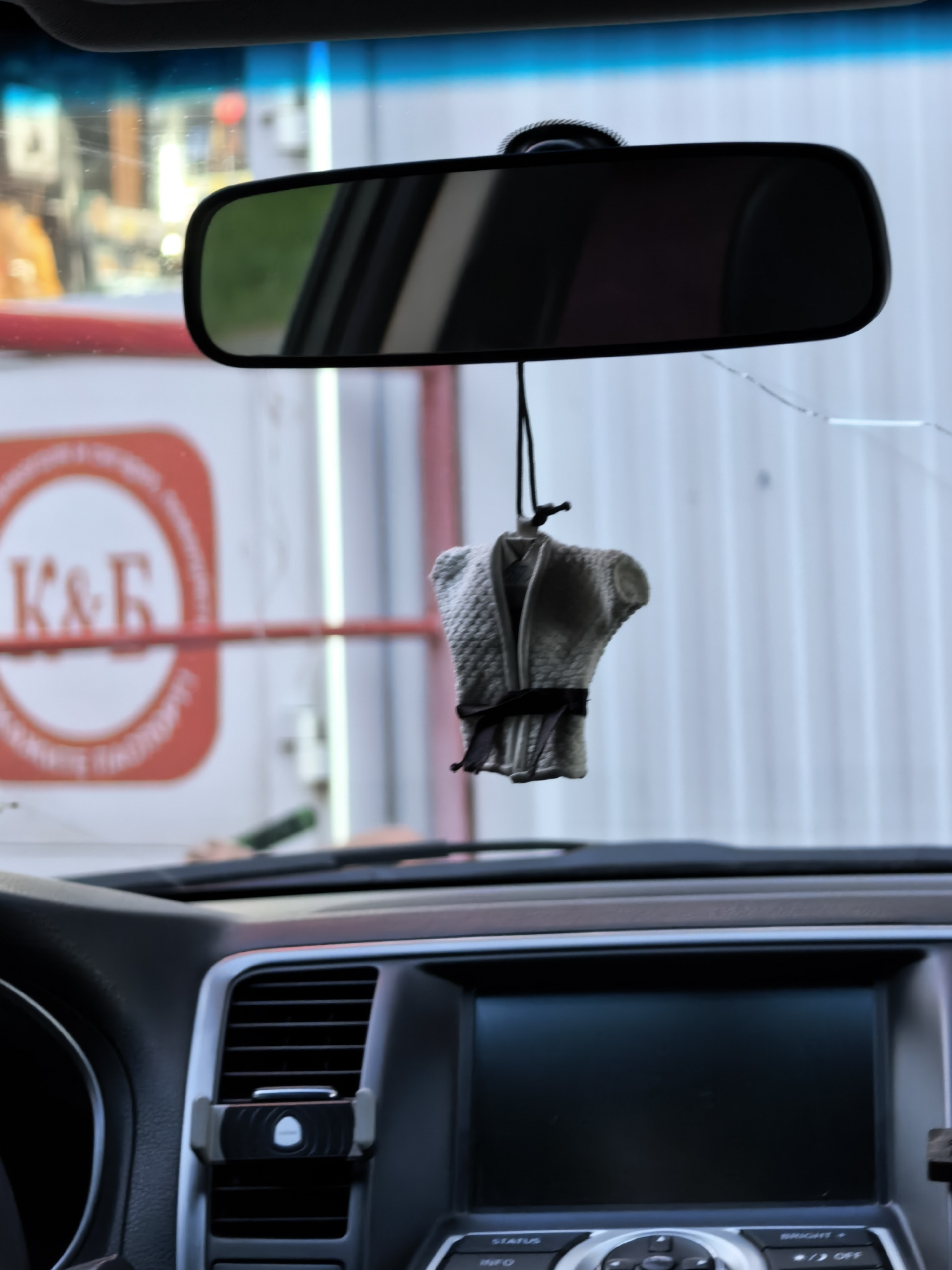 The image shows the interior of a car, specifically the dashboard and rearview mirror. Hanging from the rearview mirror is a small, fabric air freshener in the shape of a karate gi. The dashboard features a screen and various buttons. Through the windshield and reflected in the mirror, a building is visible with a sign featuring the letters "K&Б," indicating the location is Sterlitamak, Russia.