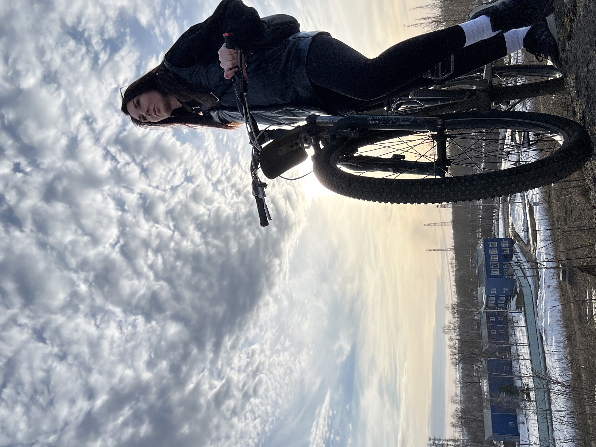 The image captures a female individual standing alongside a dark mountain bicycle on a dirt and rocky incline in Chaikovsky, Russia. The subject, viewed from a low angle, has long dark hair and wears a dark long-sleeved top, a dark vest, dark leggings, white socks, and dark athletic shoes. She grips the bicycle's handlebars with both hands and gazes upwards and to the right. The bicycle features front suspension (branded "Gila Plus") and knobby tires; a dark bag or pouch is attached to its top tube. In the middle ground, below the incline, are patches of snow, bare deciduous trees, and a light blue fence. Three distinct blue-colored buildings with white window frames and dark roofs are visible. In the far distance, several tall industrial structures, consistent with power transmission towers, rise against the horizon. The sky is dominated by extensive scattered clouds, and the low sun, positioned to the left of the bicycle, casts a warm glow and a lens flare. The presence of snow and bare trees indicates late winter or early spring conditions.