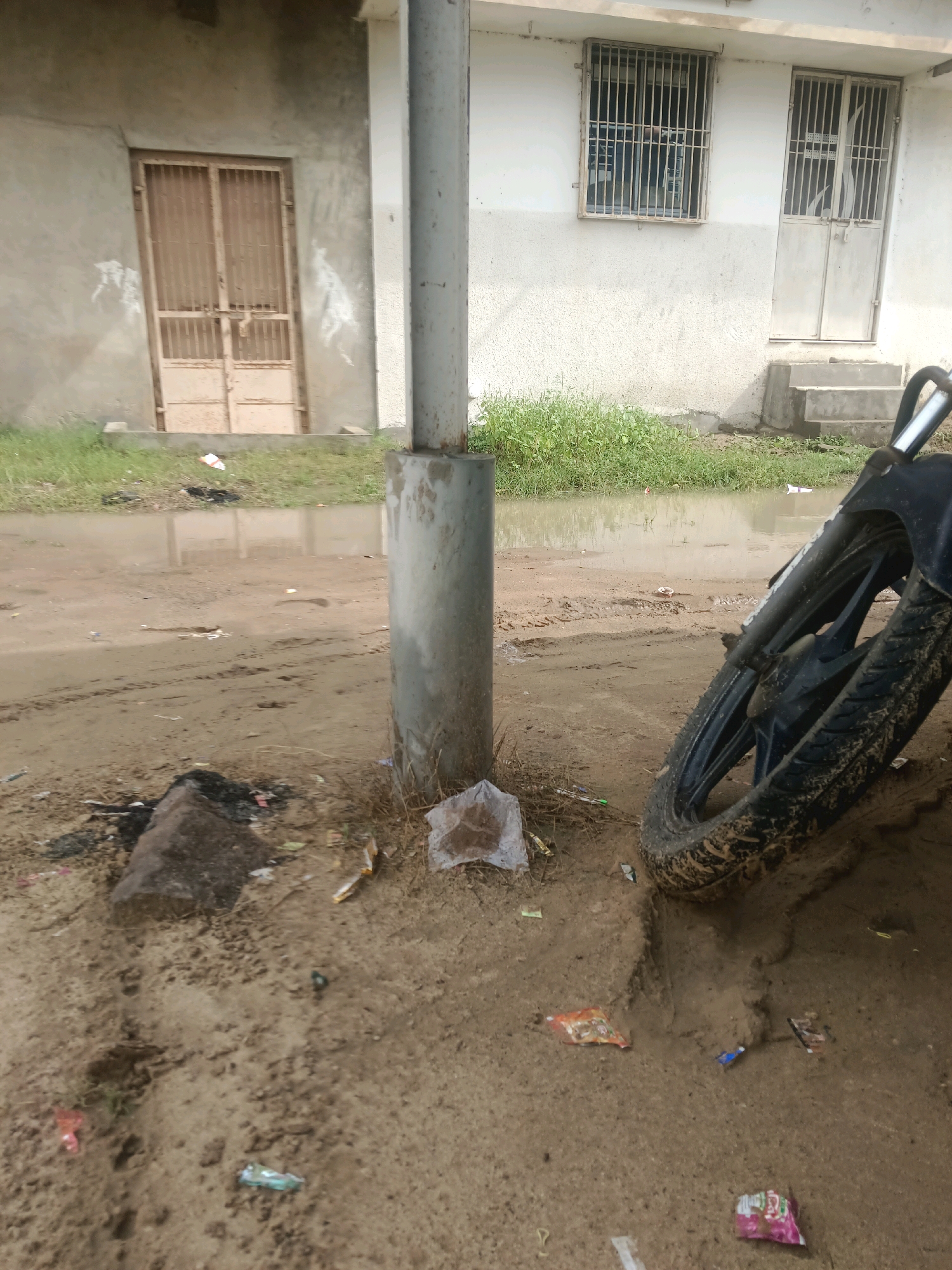 The image depicts an outdoor scene in Tokariya, India, characterized by a muddy and partially flooded dirt ground. In the foreground, the front tire and part of the frame of a dark-colored motorcycle are visible, leaning against the muddy earth. The ground is littered with various pieces of trash, including plastic wrappers and what appears to be a discarded piece of charcoal or burnt wood.
Behind the motorcycle, a concrete pillar stands prominently. Further back, there is a building with a light-colored, textured wall. The building features barred windows and metal doors. To the left, there is another building with a similar facade and a double metal door. Green grass and vegetation are present in the background, bordering a large puddle of murky water that reflects the sky. The overall impression is one of an unkempt, possibly post-rain or poorly maintained urban or semi-urban environment.