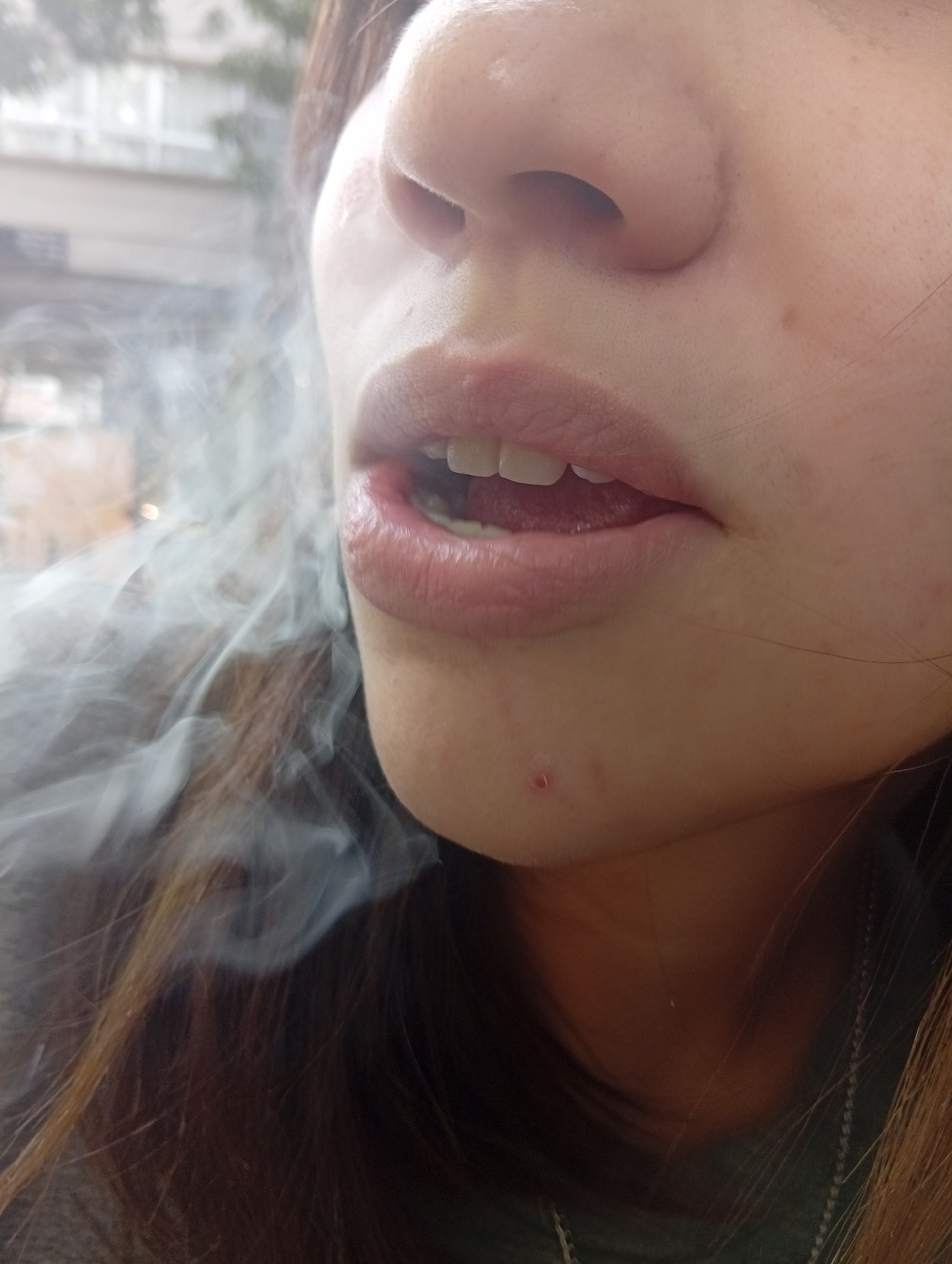 A close-up view depicts a person's face, specifically the area from the nose to the upper chest. The person's mouth is slightly open, revealing their upper teeth and a portion of their tongue. Smoke is visible emanating from the lower left side of the image, partially obscuring the left side of the face. A small blemish is noticeable on the chin. The person has brown hair. A necklace is visible on the lower right. The background is blurred, but appears to show a building through a window. The location cannot be confirmed as Buenos Aires, Argentina.