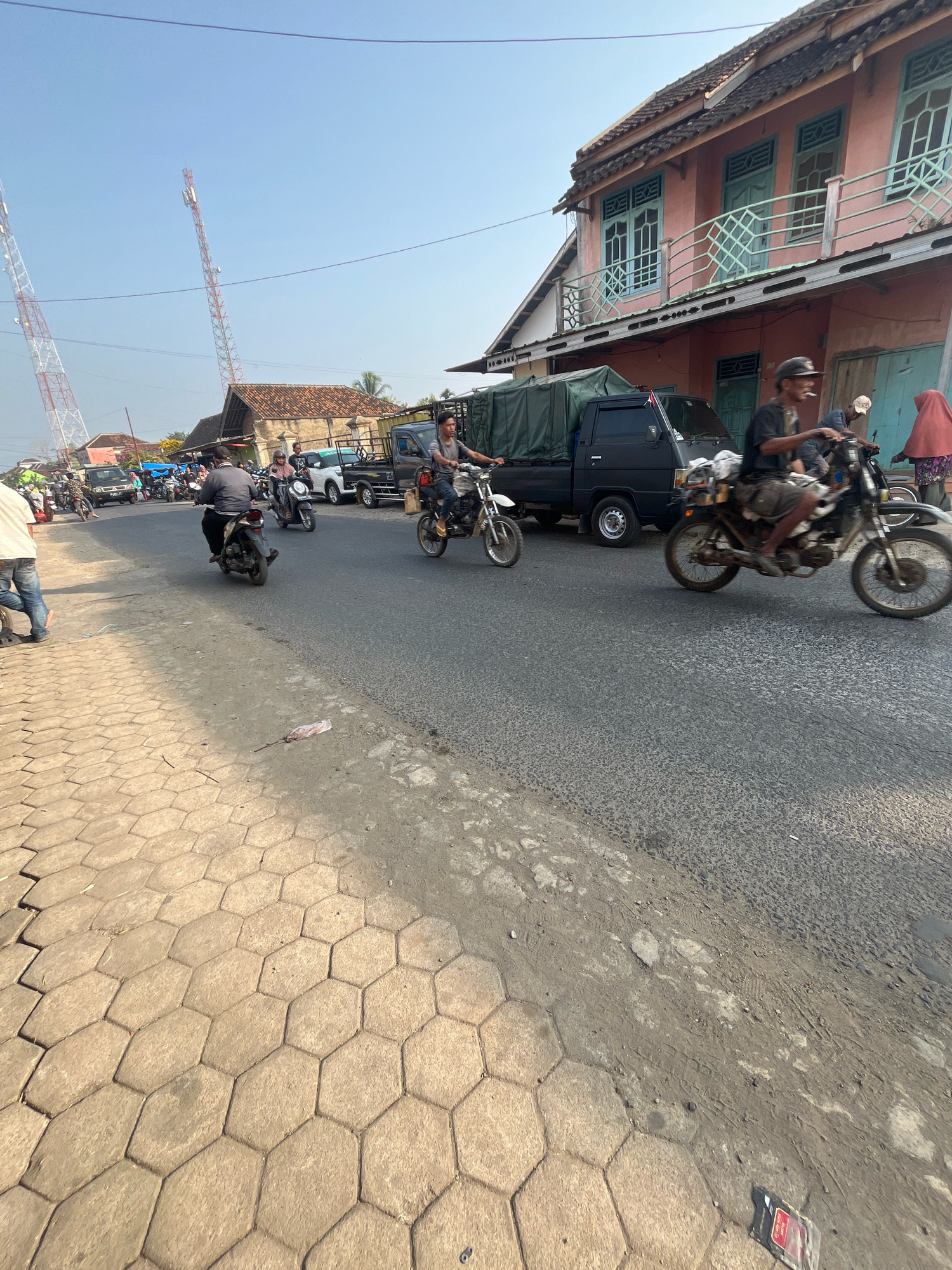 In Kemuning, Indonesia, a street scene unfolds under a clear sky. The image captures a roadside with a paved walkway of hexagonal tiles adjacent to a two-lane asphalt road. The road is active with vehicular traffic, including multiple motorcycles and a black truck.
On the right side of the road, a pink multi-story building stands with light blue framed windows and a balcony. A person wearing a red headscarf and a patterned skirt stands near the building's entrance. To the left of the building, a black truck with a green tarp covering its bed is parked.
Several individuals are riding motorcycles on the road. In the foreground on the right, a person wearing a hat and shorts is riding a motorcycle, with another motorcycle and rider partially visible behind them. Further down the road on the left, a person in a grey shirt and black pants is riding a motorcycle, with another motorcycle and rider ahead of them. More motorcycles and vehicles are visible further in the background, suggesting moderate traffic.
Two tall communication towers, one white and one red and white, are visible in the distance against the sky, indicating the presence of infrastructure in the area. The overall impression is a bustling street scene in a populated Indonesian town.
