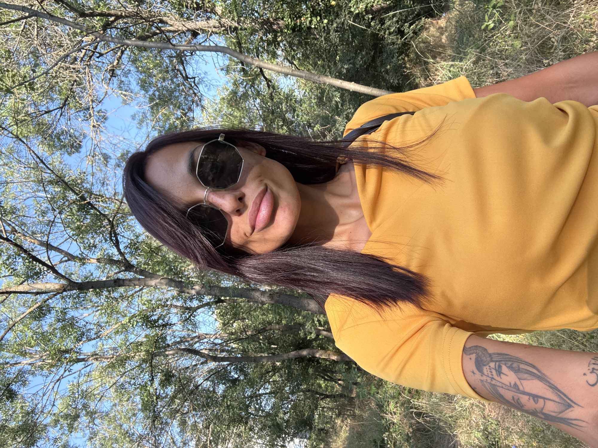 A woman with dark hair and sunglasses is taking a selfie outdoors in a wooded area with trees and foliage. She is wearing a bright yellow t-shirt. Her left arm, visible in the foreground, has a tattoo of two faces and some text. The location is identified as Pleven, Bulgaria. The lighting suggests it is daytime, with sunlight filtering through the trees.