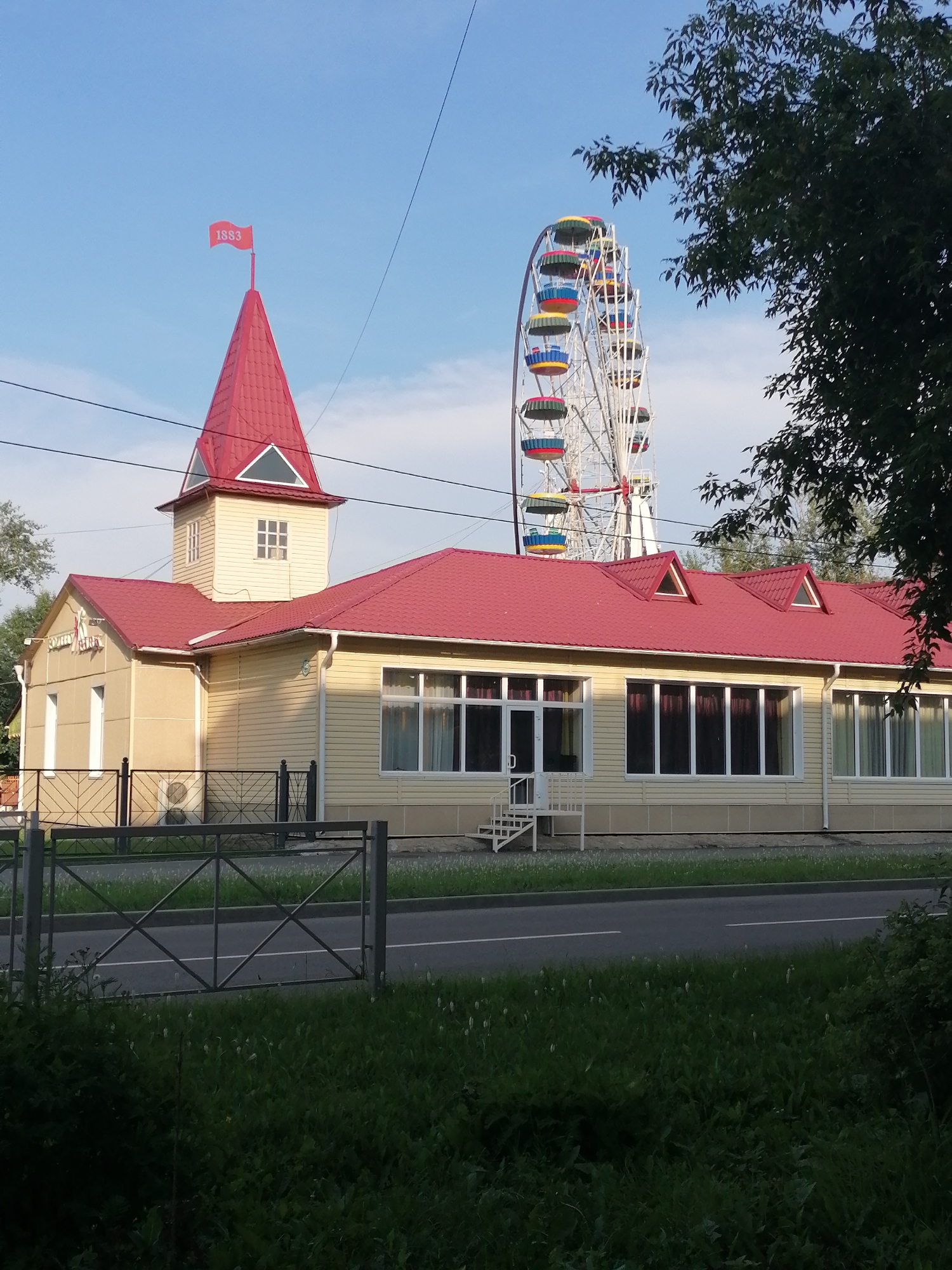 The image depicts the Ferris wheel amusement ride in the city of Shadrinsk, Russia, located in the Kurgan Oblast. The Ferris wheel, a prominent structure, features colorful cabins and is set against a clear blue sky. In the foreground, a light-colored building with a red roof is visible. The building has large windows and a distinctive pointed tower topped with a red flag displaying the year "1883". The scene is framed by green foliage and trees, suggesting an outdoor, possibly park-like setting. A metal fence runs along the front of the building, separating it from a paved road. No people are visible in the image.