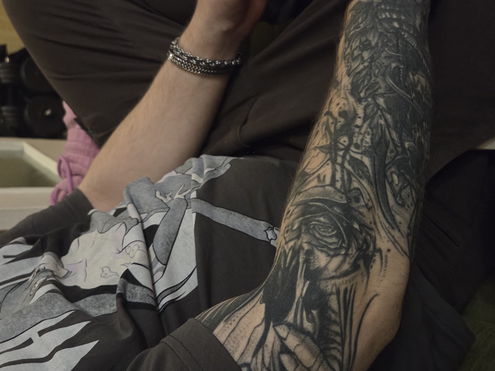 A view of an arm with a tattoo, wearing a bracelet. The tattooed arm is on top of a graphic t-shirt, which is on top of brown pants. Weights are visible in the background. The location is unknown.