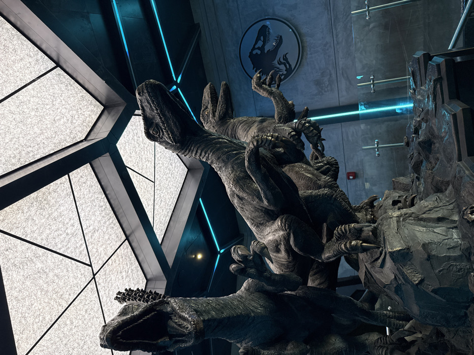 Three dark-toned Velociraptor statues are prominently displayed on a sculpted, rocky base in what appears to be an indoor themed environment. The scene is located within the Jurassic Park/World area, specifically in the queue area for the Jurassic World VelociCoaster at Universal's Islands of Adventure in Orlando, Florida, United States of America.
The statue on the far left features a raptor with its mouth agape, head tilted upward. A central raptor is positioned with its head forward and slightly raised, claws prominently extended from its forelimbs. A third raptor is partially visible behind and to the right of the central figure, also in an active stance. The raptors are detailed, showing musculature and texture.
Above, a geometrically patterned ceiling composed of translucent, illuminated panels is supported by dark structural beams. Blue linear LED lighting strips are integrated into the upper wall edges and vertical structures, contributing to the ambient illumination. On the right-hand wall, a circular black and white Jurassic Park/World logo, depicting a Tyrannosaurus Rex silhouette, is affixed. Below this logo, a red fire alarm pull station is visible on the light gray, textured wall. Portions of metal barrier railings are discernible in the lower right foreground, suggesting a public area or queue line. The overall lighting is dim, emphasizing the blue accents and the illuminated ceiling. No individuals are present in the immediate foreground.