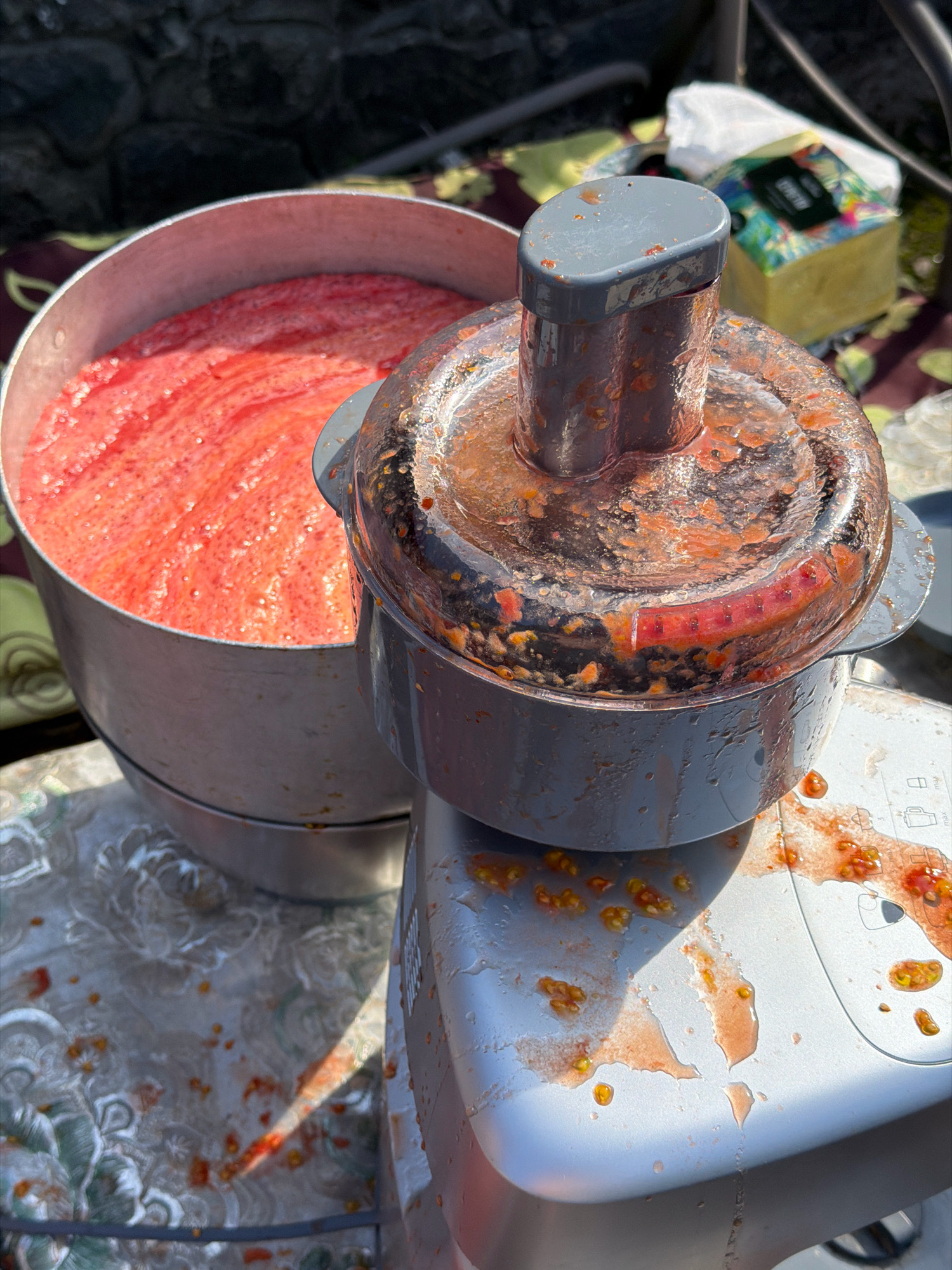 The image depicts an outdoor scene in Novosilka, Ukraine, with a food processor and a large metal pot filled with pureed tomatoes. The food processor is smeared with tomato puree and seeds, indicating recent use. Tomato pulp and seeds are also scattered on the table and the ground around the appliances. In the background, a partially visible roll of paper towels sits on a patterned tablecloth. The overall impression is of a food preparation process taking place outdoors.