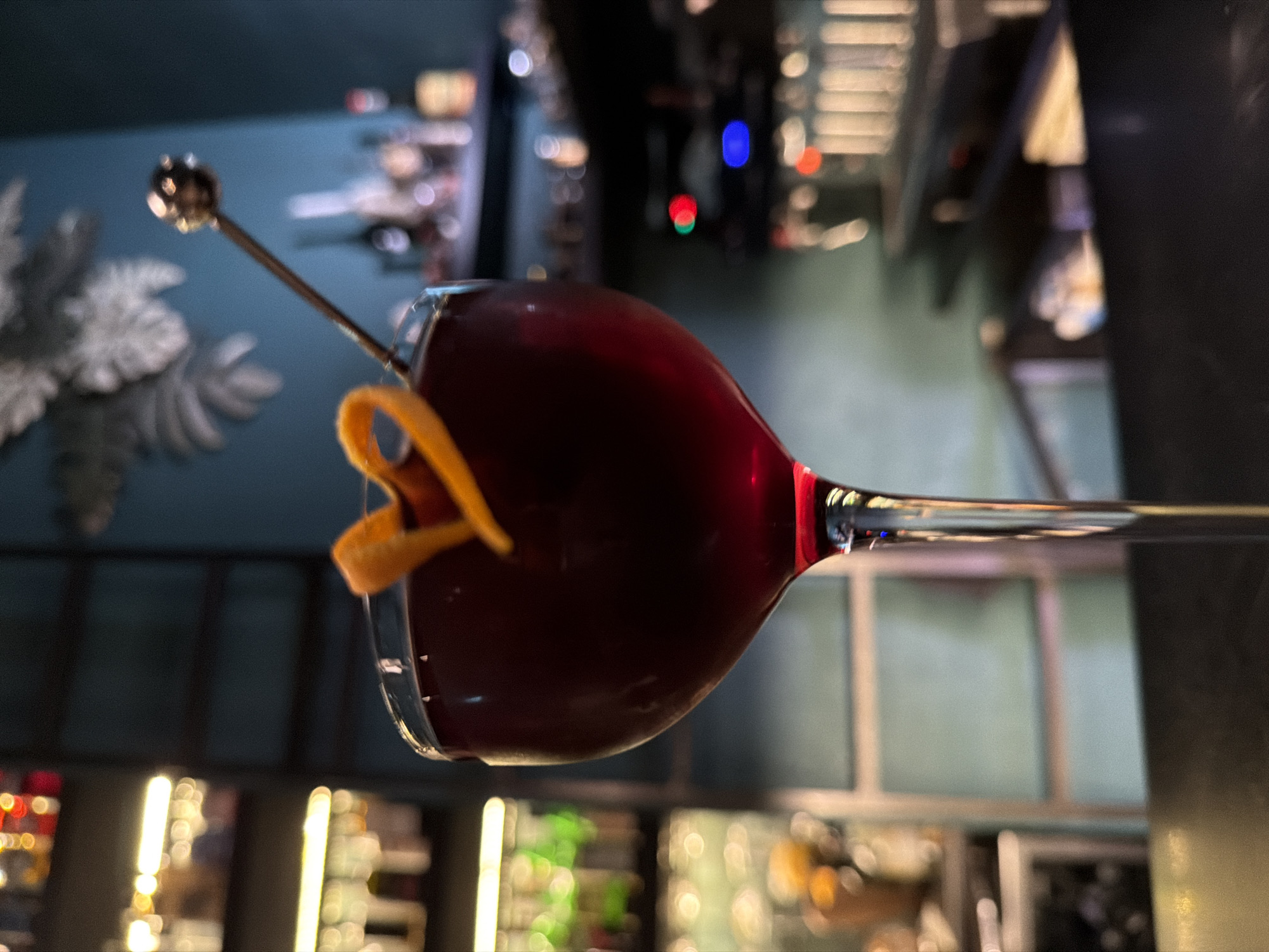 The image displays a deep red or burgundy cocktail served in an elegant, clear stem glass. The glass features a long, slender stem and a rounded bowl filled with the dark-hued beverage. A decorative garnish, an orange peel twisted into an artistic shape, hangs over the rim of the glass. A thin, metallic cocktail stirrer or pick, topped with a small, shiny spherical detail, is submerged into the drink.
The background is heavily blurred, creating a soft bokeh effect with various lights and indistinct shapes, suggesting an indoor setting, likely a bar or lounge. On the left, a dark blue or teal wall is visible, adorned with a white, leaf-like decorative element that is also out of focus. On the right and in the lower portion of the background, numerous blurred lights in shades of red, blue, green, and yellow/white hint at bottles on shelves, bar equipment, or ambient lighting. The scene suggests an evening or nighttime setting due to the low light and prominent artificial illumination. No people or visible text are present.