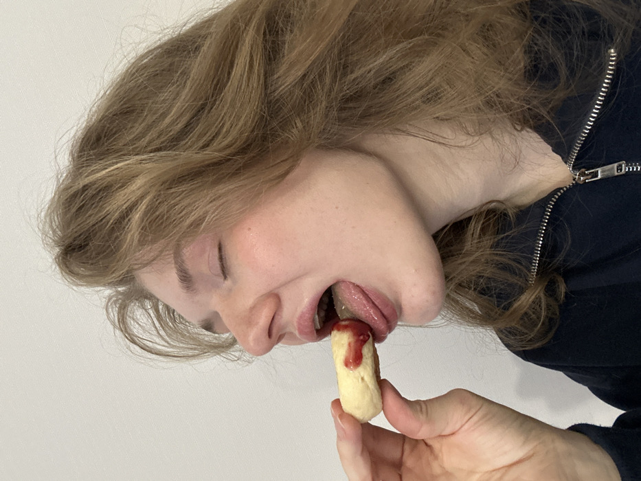 A fair-skinned person with wavy, light brown hair, eyes closed, and head tilted, is captured from a low-angle perspective. The individual is in the act of eating. The tongue is extended, coming into contact with a small, oblong, beige-colored pastry. A red sauce, possibly jam or ketchup, is drizzled on top of the pastry. The pastry is being held by a hand with fair skin. The person is wearing a dark blue zippered sweatshirt. The background appears to be a plain white surface.