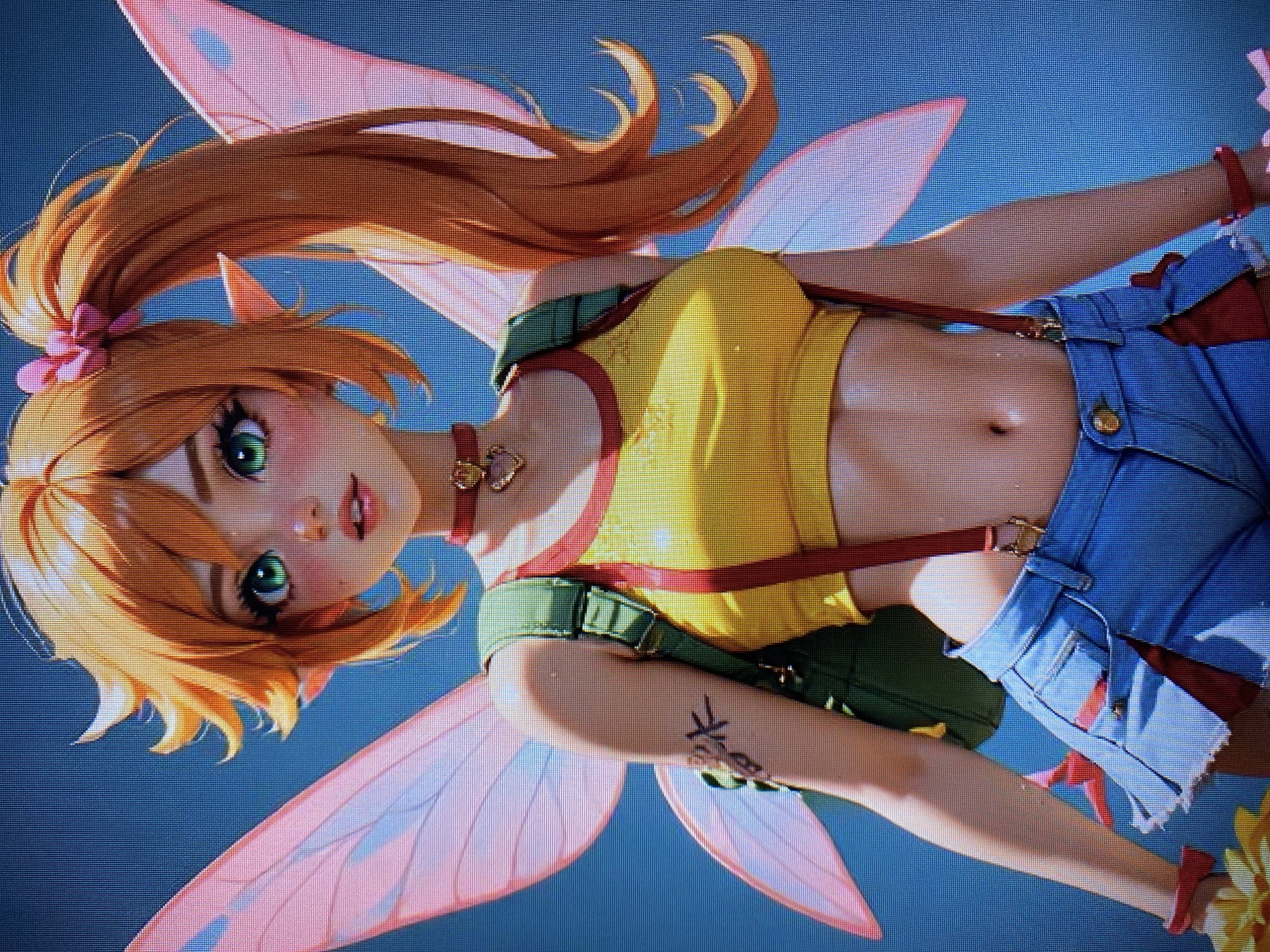 The media file shows a 3D rendered image of a young woman with orange hair, green eyes, and translucent pink fairy wings. She is wearing a yellow crop top with red trim, blue denim shorts with red suspenders, and a red choker necklace with a heart-shaped pendant. Her hair is styled in a high ponytail with a pink bow. She has a small tattoo on her left arm. The background is a solid blue. The image is a close-up shot, focusing on the character's upper body and face. The lighting suggests a bright, possibly daytime scene. There are no discernible location cues, text, or specific events indicated in the image. The character's expression is neutral to slightly curious.