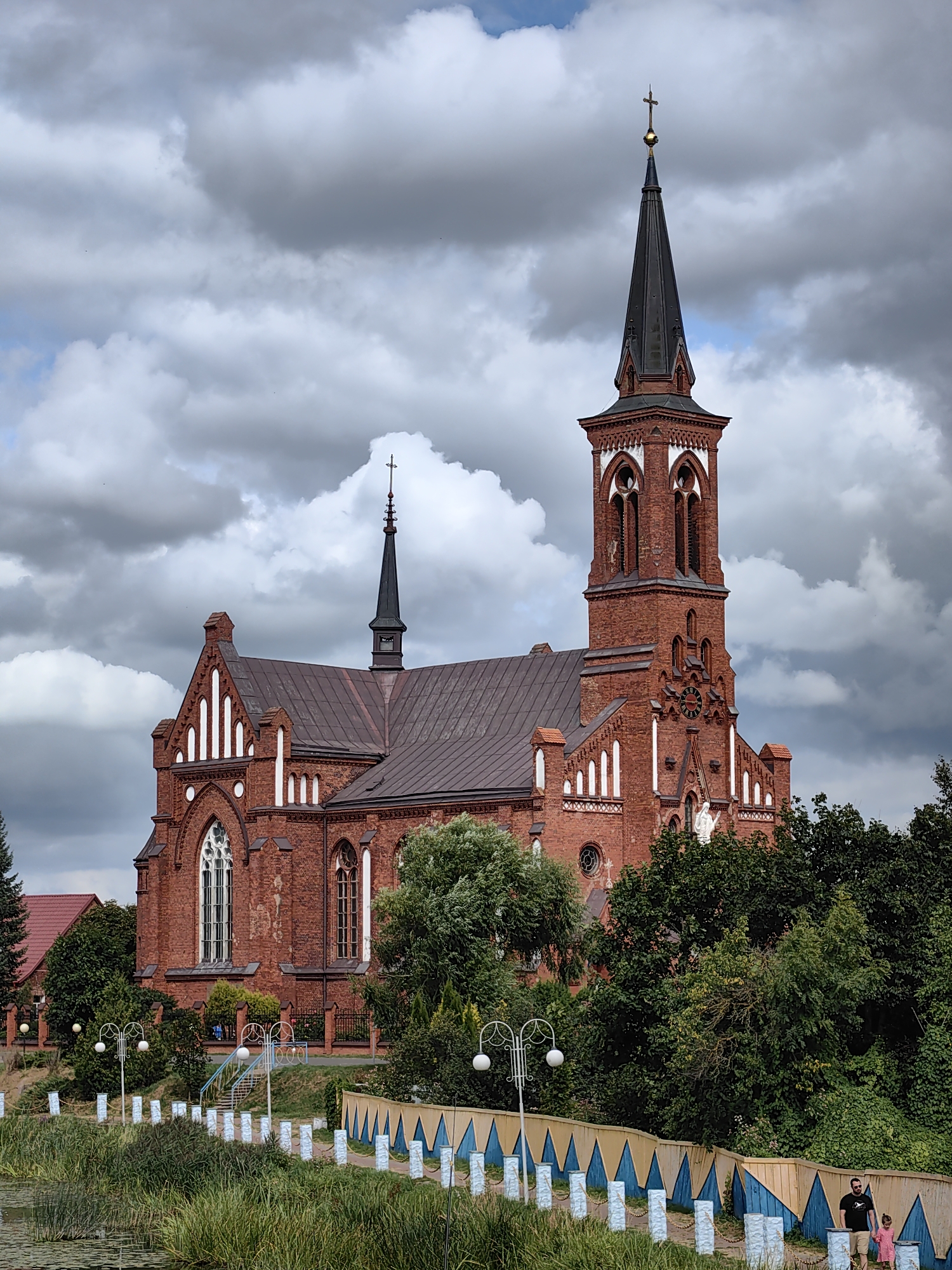 The image shows a red brick church with a tall, dark spire topped with a cross against a dramatic cloudy sky. The church has Gothic architectural elements, including pointed arches and intricate brickwork. In the foreground, there is a body of water with tall grass along its edge. A decorative fence with blue and yellow triangular patterns runs along the bank, with white posts. A man and a young girl are walking up a slight incline on a path near the fence. The scene appears to be located in the city of Pastavy, Belarus, and the church is likely the Church of the Transfiguration.