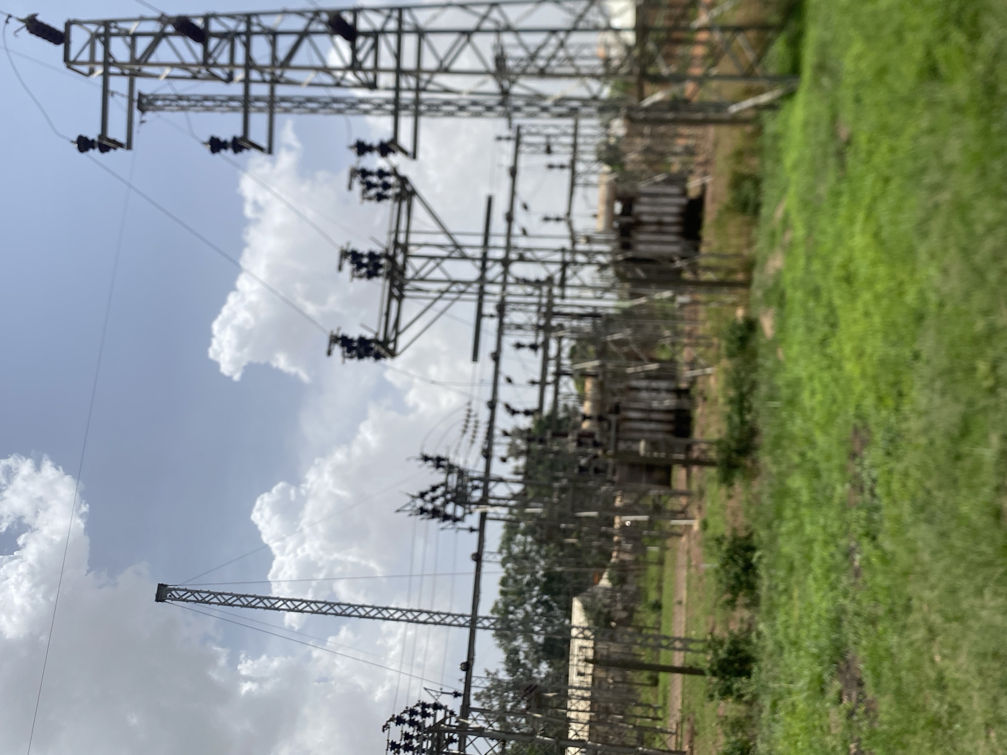 The image shows a power distribution station in Bukuru, Nigeria. The station contains tall, metal lattice towers supporting high-voltage power lines and electrical equipment. Multiple transformers are visible. The ground around the station is covered in green grass and vegetation. The sky is partly cloudy.