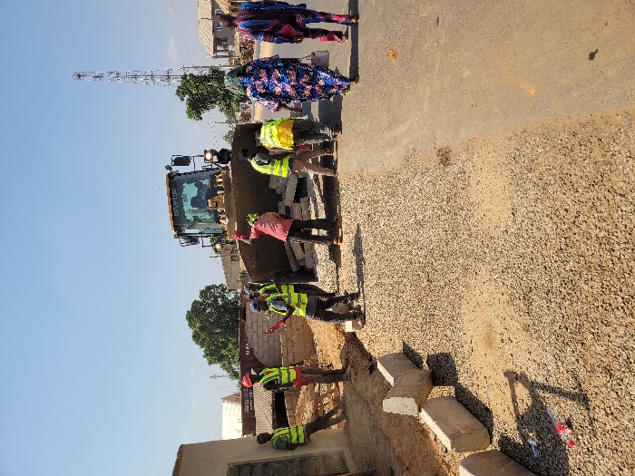 The image depicts a daytime road construction scene in Ube, Nigeria, under a clear blue sky. In the midground, a large yellow Caterpillar bulldozer is positioned with its front blade lowered, containing several white blocks. Six male workers are present around the bulldozer and the immediate construction area, mostly wearing high-visibility vests (yellow or green) and some with hard hats. One worker in a red shirt and red hard hat stands near the bulldozer's blade, with his right arm raised. Another worker in a blue cap and blue vest holds a red object. One worker leans against a light-colored building wall on the far left.
The foreground on the left features a section of ground covered with light brown gravel, bordered by several concrete curbstones, some upright and some toppled. To the right, an existing darker asphalt or dirt road surface is visible. Two women, wearing colorful patterned traditional clothing, walk away from the camera on the right side of this road.
The background includes various low-rise buildings with corrugated roofs, scattered green trees, and a tall, slender cellular communication tower prominently positioned on the right side. The overall scene suggests ongoing infrastructure development.
