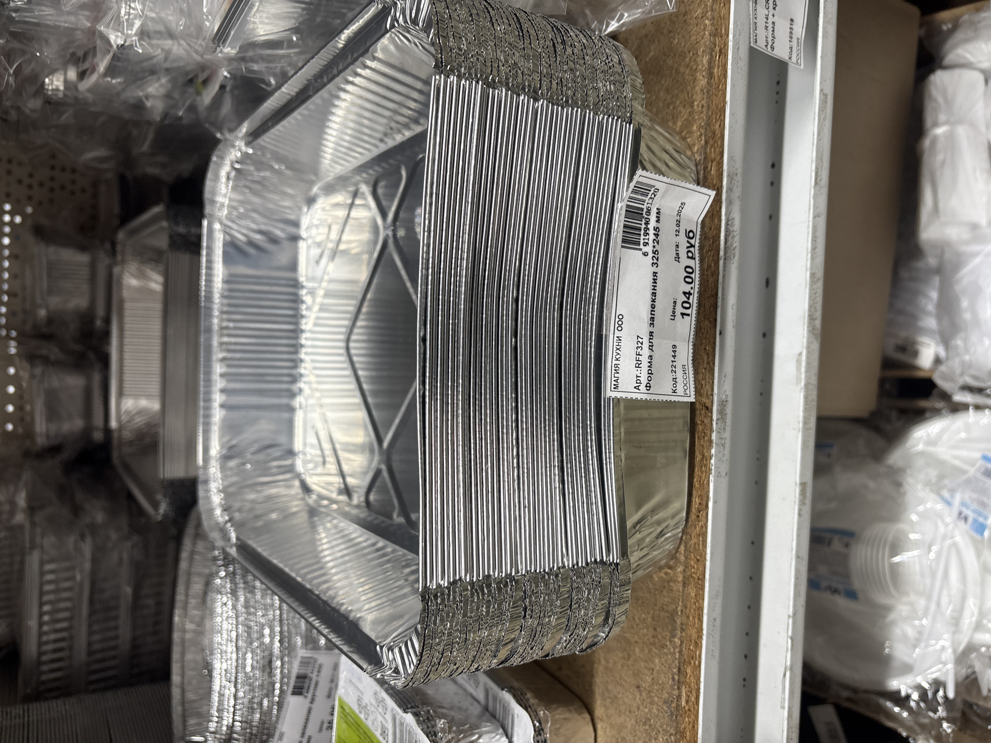 The image shows a stack of square aluminum baking pans for sale. A price tag affixed to the stack indicates that they cost 104.00 rubles. The tag also includes the following information: "MAGIC KITCHEN LLC," article number "RFF327," description "Baking dish 325*245 mm," code "221449," and the country of origin, "Russia." The date on the label is 12.02.2025. Other items in plastic packaging are visible in the background, suggesting the pans are located in a retail environment. The location is likely Krasnoyarsk, Russia, as the label mentions the country of origin as Russia.