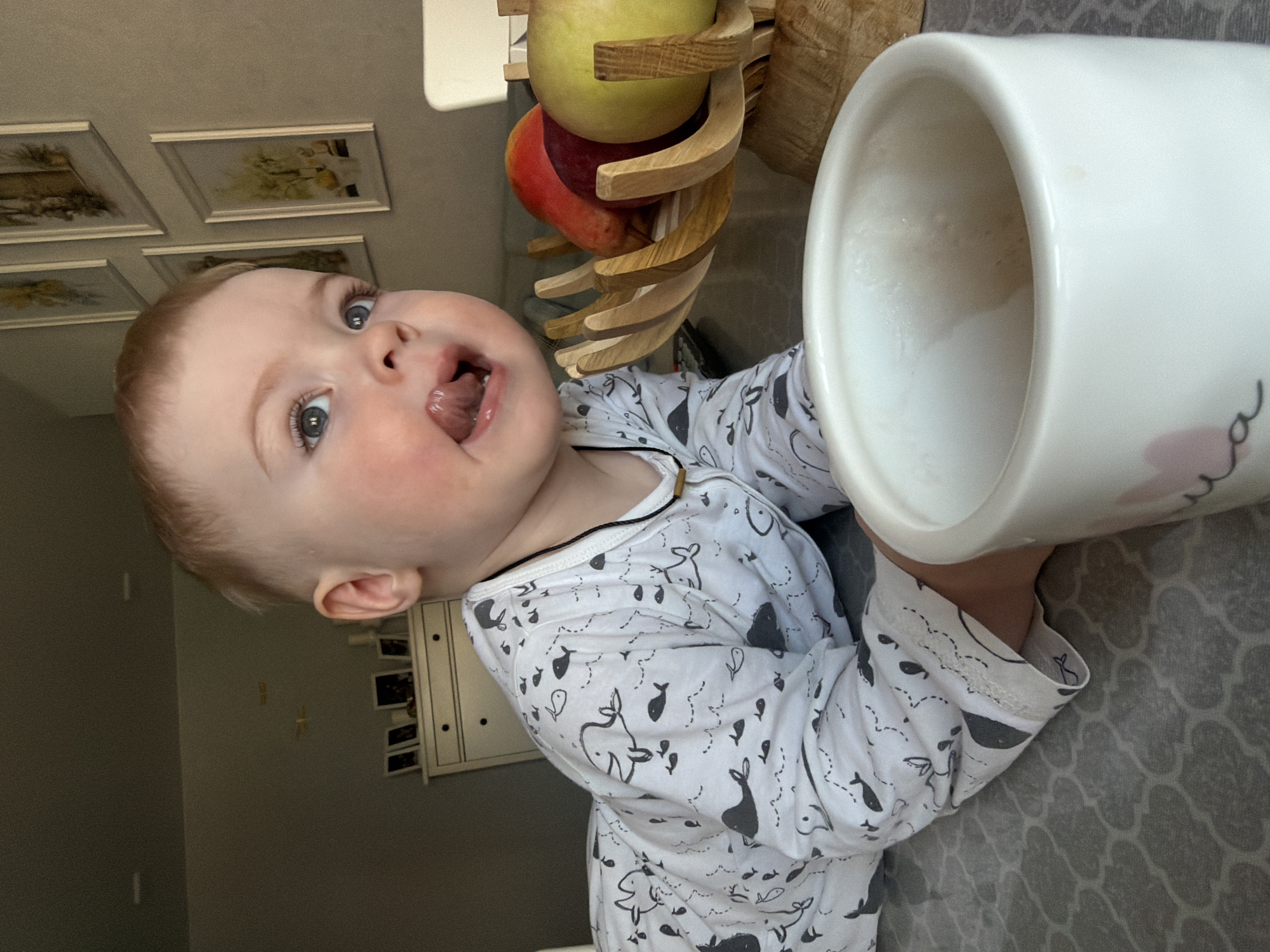 A toddler in a white pajama with grey whale patterns is looking up at the camera with an open mouth. The background contains a white wall with four framed pictures, a wooden fruit stand with a red apple and a yellow pear, and a white mug filled with a light-colored liquid. A grey patterned surface is beneath the child and mug. The location cannot be determined within Votkinsk, Russia.