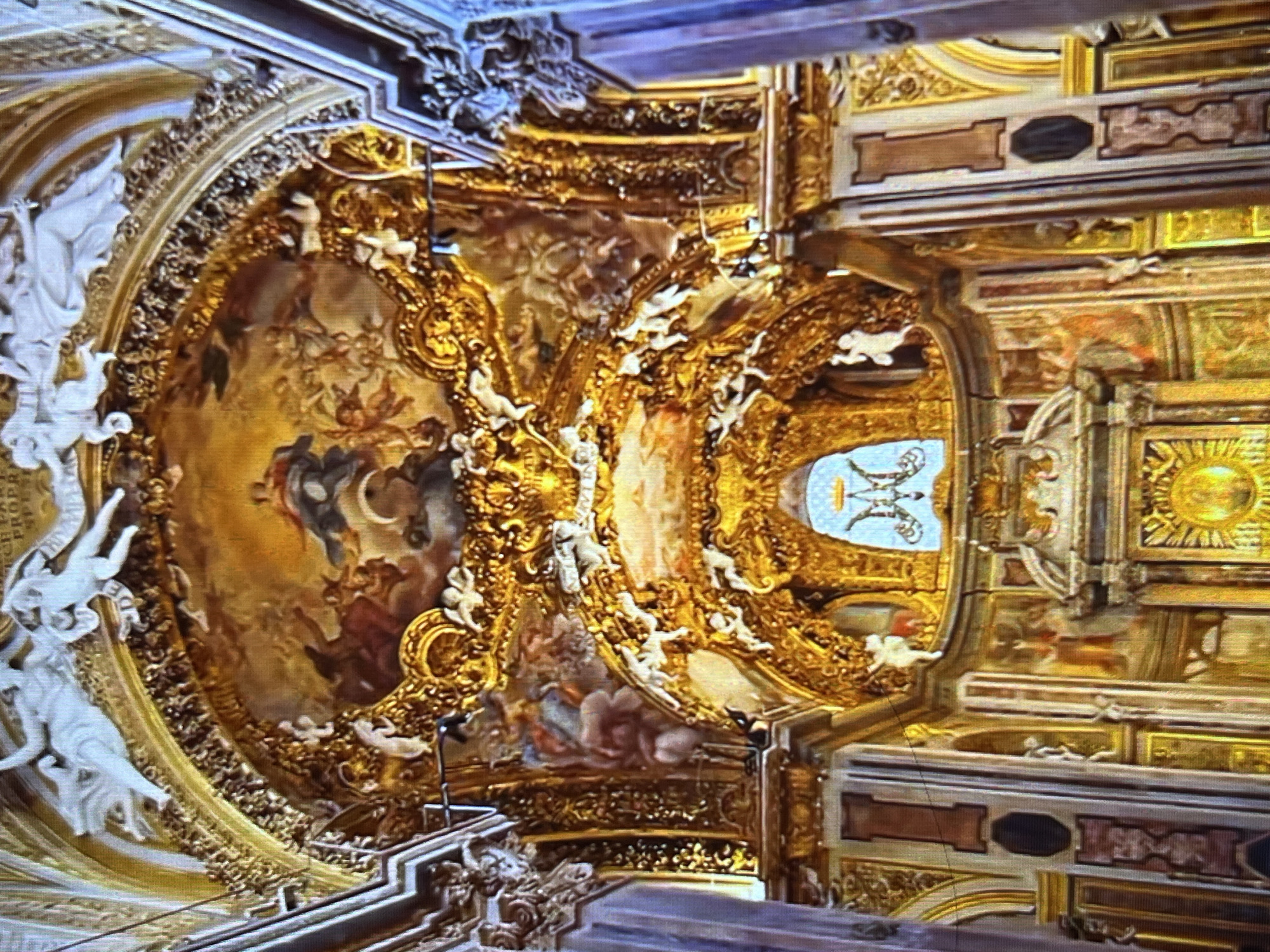 The image captures an ornately decorated ceiling, likely within a church or similar religious structure. The ceiling features intricate gold detailing, with paintings and sculpted figures. A central, oval-shaped painting depicts a scene with human figures and celestial elements. The surrounding architectural elements include decorative moldings, panels, and possibly a coat of arms. Given that Tinaquillo is a city in Venezuela, the location is likely a church or historic building within that city. It appears to be an interior shot from underneath, viewing upward at the elaborate ceiling.