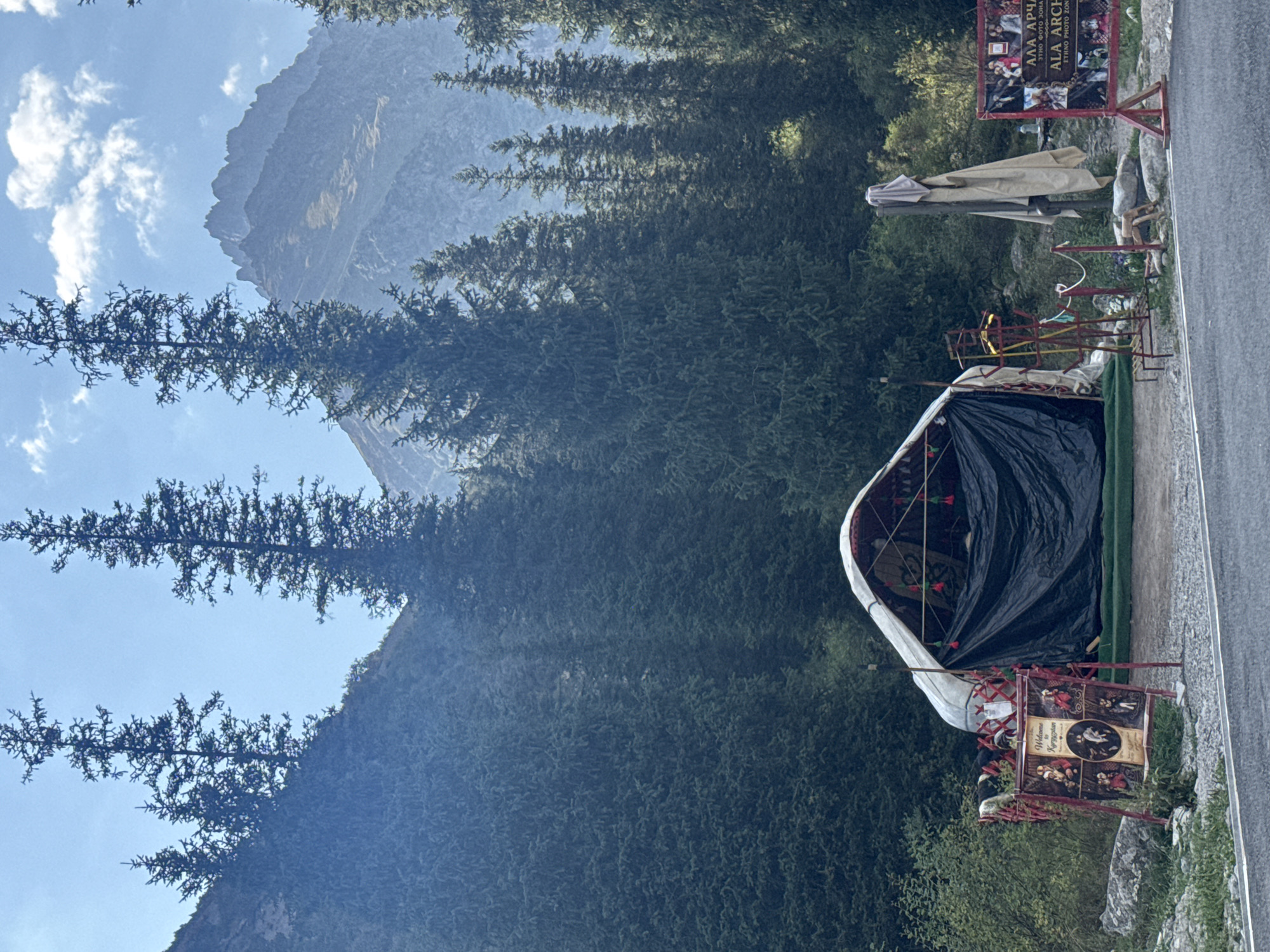 In an outdoor, mountainous setting with tall evergreen trees and hazy peaks in the background, a traditional yurt with a white canvas roof and black tarp covering the entrance stands on a gravel ground. A sign with the text "Welcome to Kyrgyzstan" and several circular images is displayed to the left of the yurt. To the right, another sign with Cyrillic text and images is visible. A clothesline with a draped cloth hangs near the yurt, and a decorative metal structure with colorful elements is positioned in front of it. The scene appears to be a tourist attraction or cultural display in the Ala Archa National Park in Kyrgyzstan.