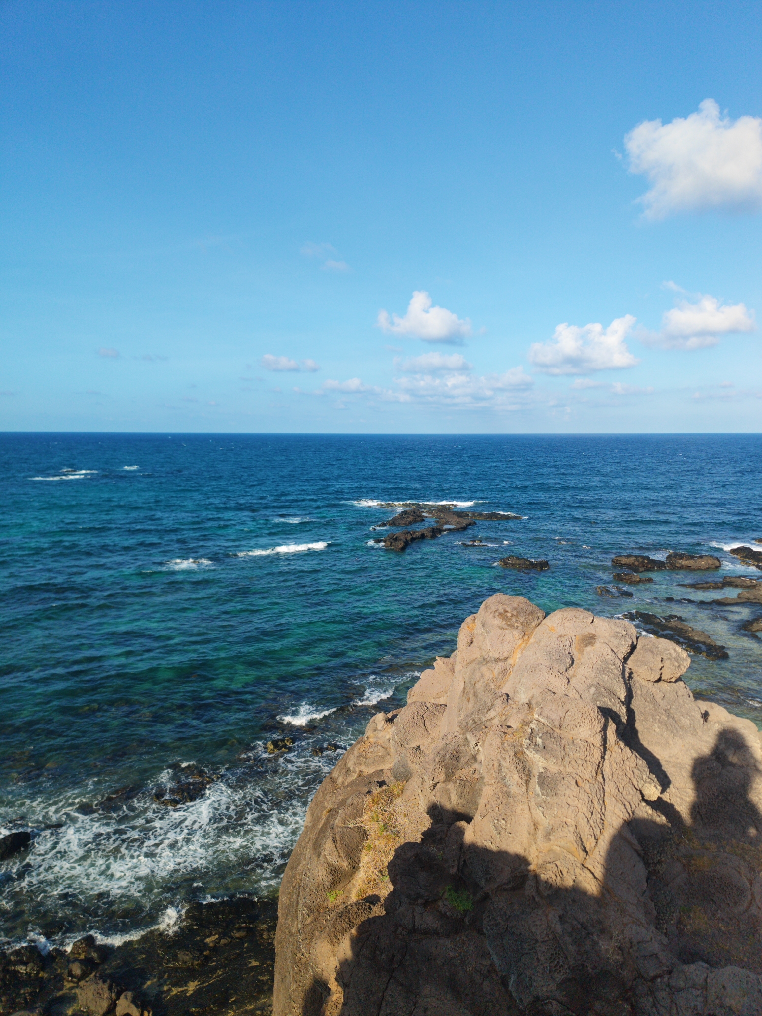 The image captures a coastal scene in Phú Quý Special Administrative Region, Vietnam. The scene features a rocky coastline with a prominent, tan-colored rock formation in the foreground. The ocean stretches towards the horizon, displaying shades of blue and turquoise. Scattered rocks are visible in the water near the shore. The sky is blue with a few white clouds. The shadows of people are cast on the rock formation, indicating their presence and perspective. The overall environment suggests a natural, coastal setting.