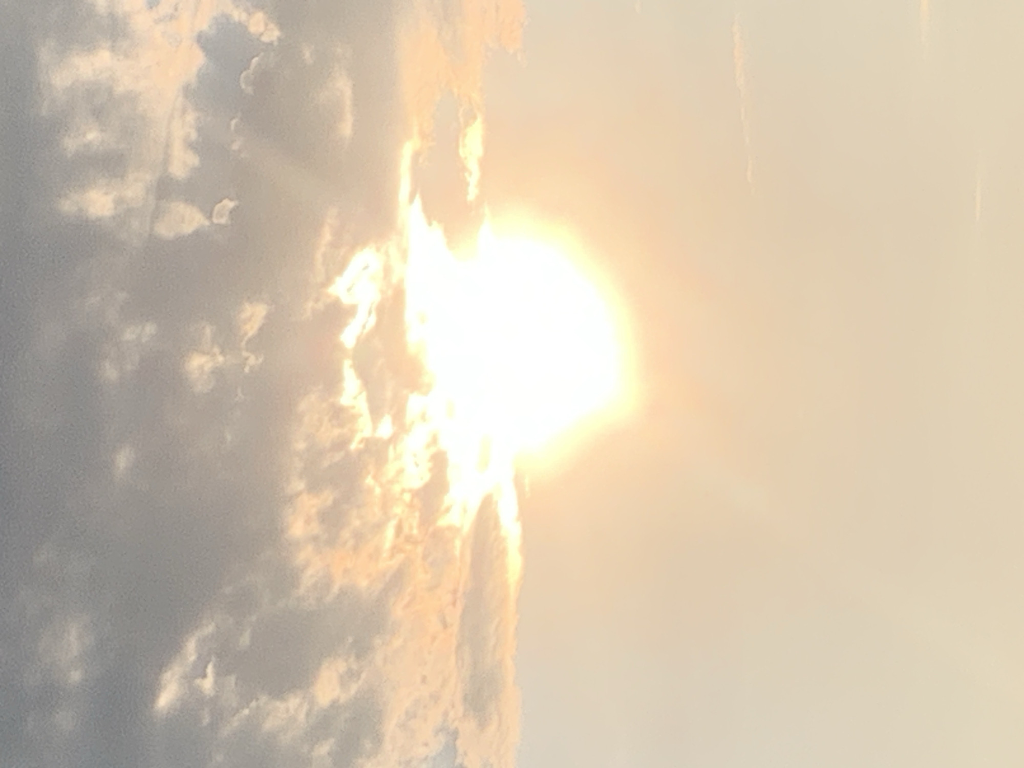 The image is a photograph of the sky, likely taken at sunset or sunrise, with the sun prominently visible as a bright, luminous orb in the center of the frame. The sun is partially obscured by clouds, which are illuminated with warm, orange and pink hues from the sunlight. The sky transitions from a pale yellow or beige at the bottom to a soft gray or blue at the top. Wispy clouds are scattered throughout the scene, with some appearing more defined than others. There are no visible objects, people, or structures in the image, as it focuses solely on the atmospheric conditions. The location in the city of Kolomyia, Ukraine, cannot be determined from the visual information provided in the image.