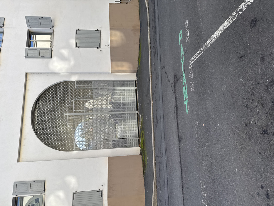 This image captures a street-level view of a building facade and adjacent asphalt road in Le Puy-en-Velay, France. The building presents a white upper section and a beige lower foundation. Central to the facade is a large arched entrance, secured by a partially lowered metal roll-down grille and a double-leaf metal gate with a square grid pattern. Four windows with light-gray wooden shutters are visible: two on the upper left are closed, one on the upper right is open revealing a reflective pane, and one on the lower right is closed. A small amount of moss grows on the sidewalk at the base of the arched entrance. The foreground consists of an asphalt road with visible cracks. Green stenciled text reading "PAYSAGE" is visible on the asphalt, along with a faded white parking line marking. A concrete curb delineates the sidewalk from the road. No individuals, vehicles, or animals are present. Distinct shadows are cast on the building's white facade, indicating directional sunlight.