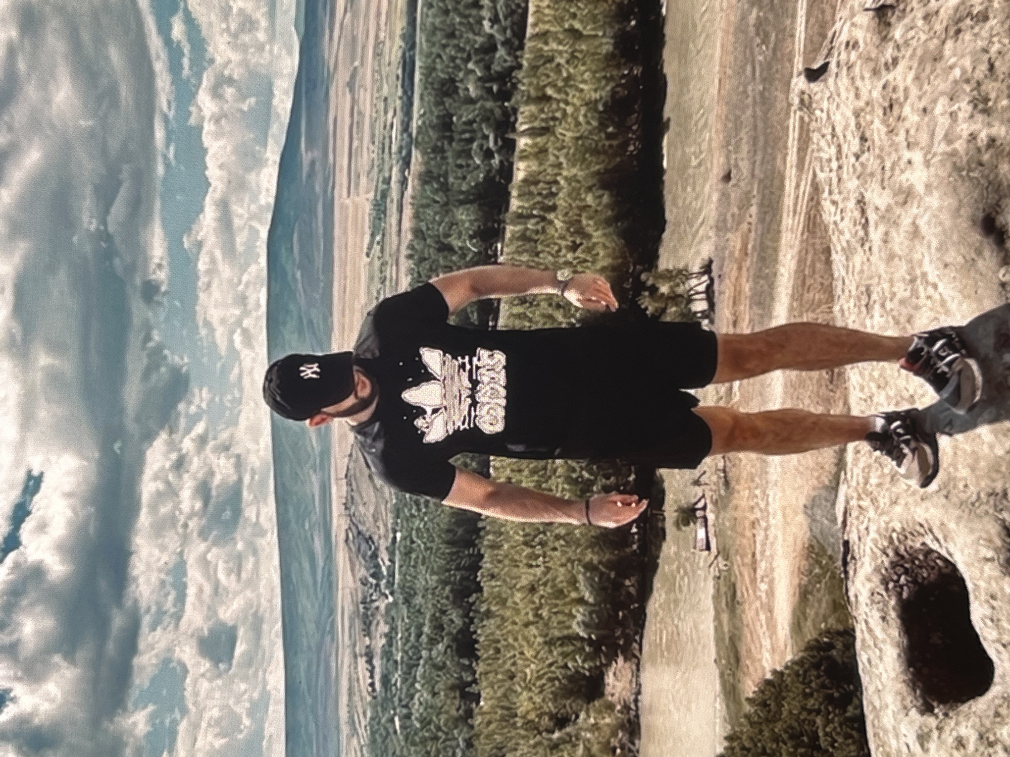 An outdoor, medium shot shows a Caucasian man standing on a rocky surface, facing away from the camera and looking down. He is wearing a black t-shirt with a white Adidas logo, black shorts, a black baseball cap with a white logo, and athletic shoes. His right arm is bent at the elbow, and he has a watch on his left wrist. The man stands on a rocky outcrop with a dark hole in the foreground. Behind him, a landscape unfolds with a river, a dense forest of green trees, and rolling hills in the distance under a cloudy sky.