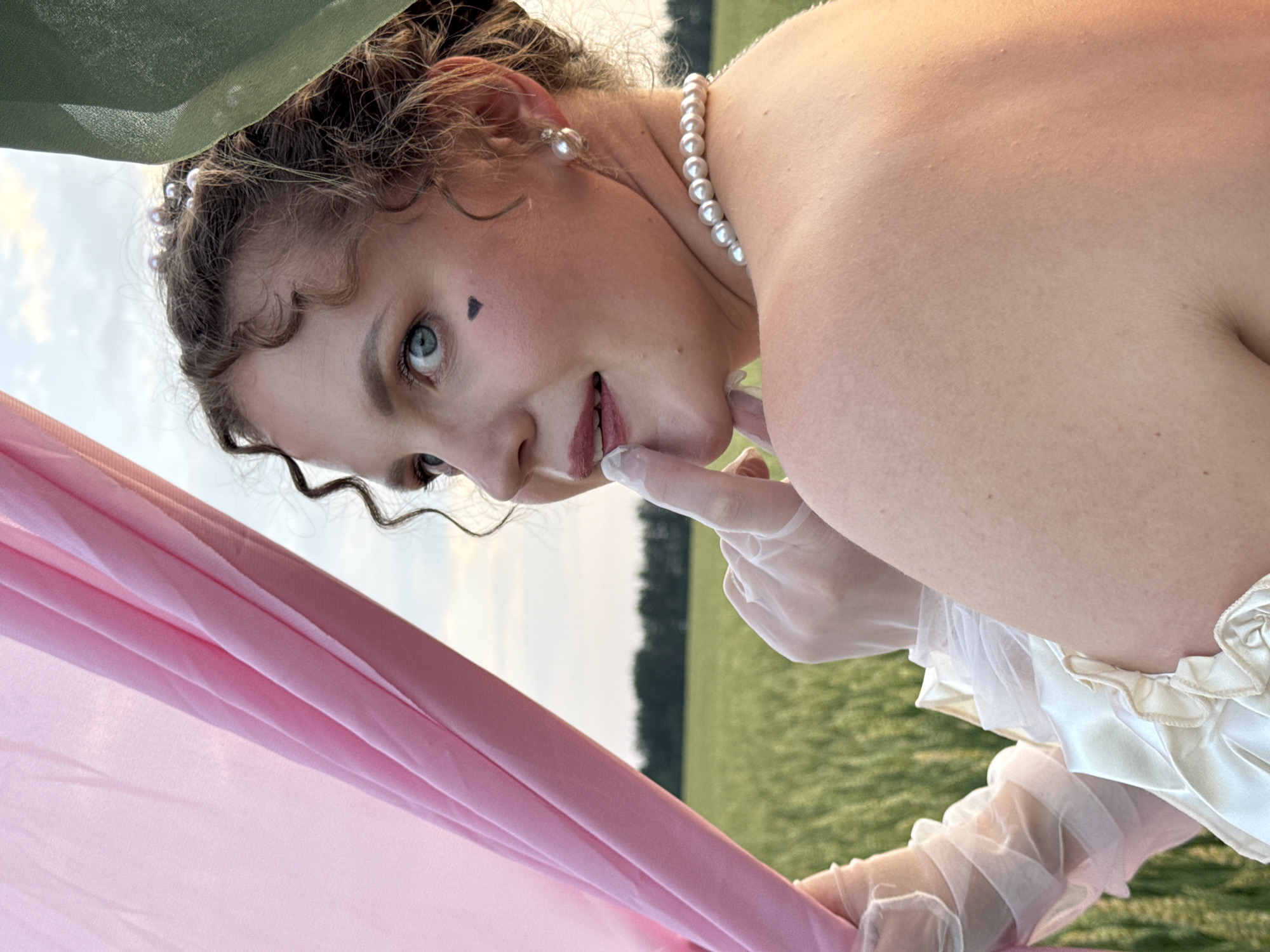 A young Caucasian woman with curly brown hair is in a field of wheat, partially obscured by sheer pink and green fabric. She is looking over her shoulder towards the camera, with her right index finger touching her lips. She has a small black heart drawn on her left cheek, and she is wearing pearl earrings and a pearl necklace. Her dress appears to be made of white satin with ruffled sleeves. The sky is a pale blue with scattered clouds, and the sun is setting, casting a warm glow on the scene. The location is Foki, Russia.