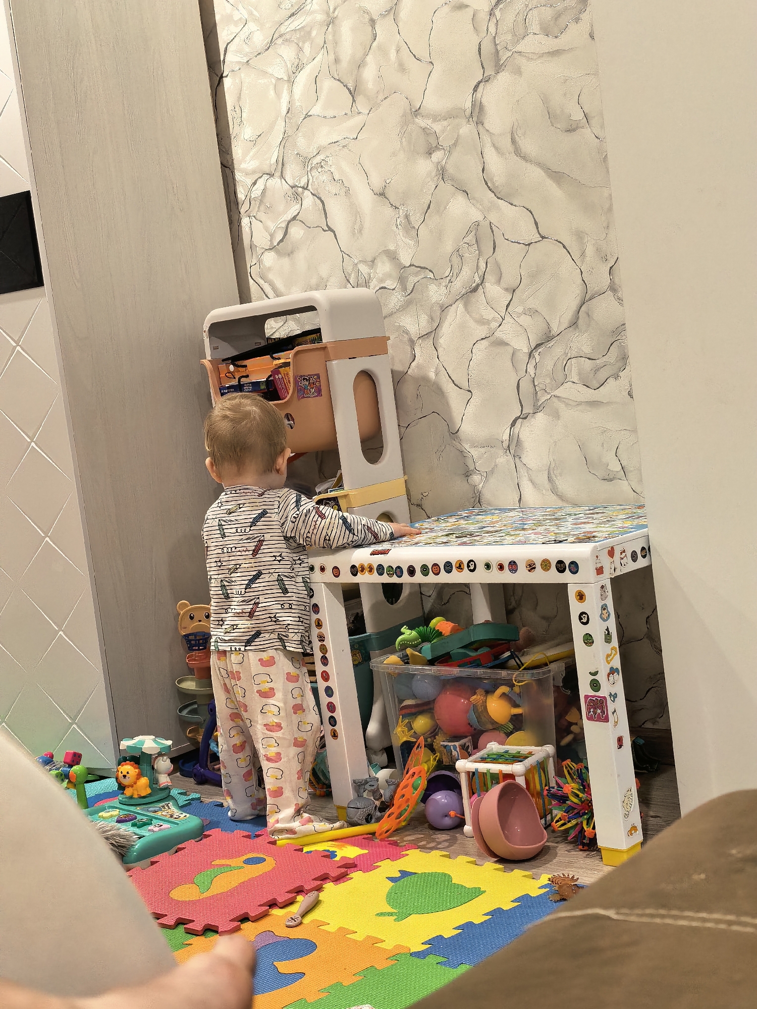 A child stands facing a colorful toy table in a room filled with toys. The child wears a striped long-sleeved shirt and patterned pajamas. The table is covered in stickers and puzzles. Various toys such as balls, containers, and interactive playthings are scattered around the floor and stored underneath the table. The floor is covered with colorful interlocking foam mats. The room's wall has a marble-like pattern, and there is a cabinet or a door to the left. The location is Unknown, Unknown.