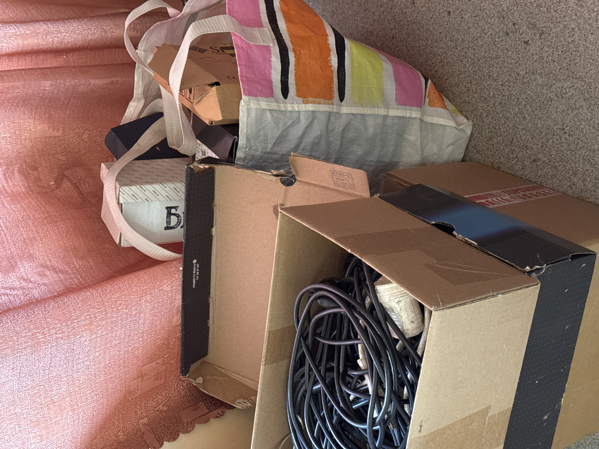 The image depicts an indoor scene, likely a storage area or a room undergoing tidying, with several cardboard boxes and a colorful tote bag filled with items. In the foreground, a large cardboard box is open, revealing a tangled mass of black electrical cords and at least one white plug or adapter. To the right of this box, another open cardboard box contains more miscellaneous items, including smaller cardboard pieces, and a glimpse of a striped fabric bag. Further back and to the right, a large, colorful tote bag with horizontal stripes of pink, orange, yellow, and black, is visible, also containing various objects. Behind the boxes and bag, a textured, pinkish-red fabric, possibly a curtain or draped material, hangs in the background. The floor appears to be carpeted with a speckled grey and beige pattern. The overall impression is one of accumulated belongings. There are no people visible in the image, and no specific landmarks or contextual clues suggest the city of Naro-Fominsk, Russia, or any other specific location.