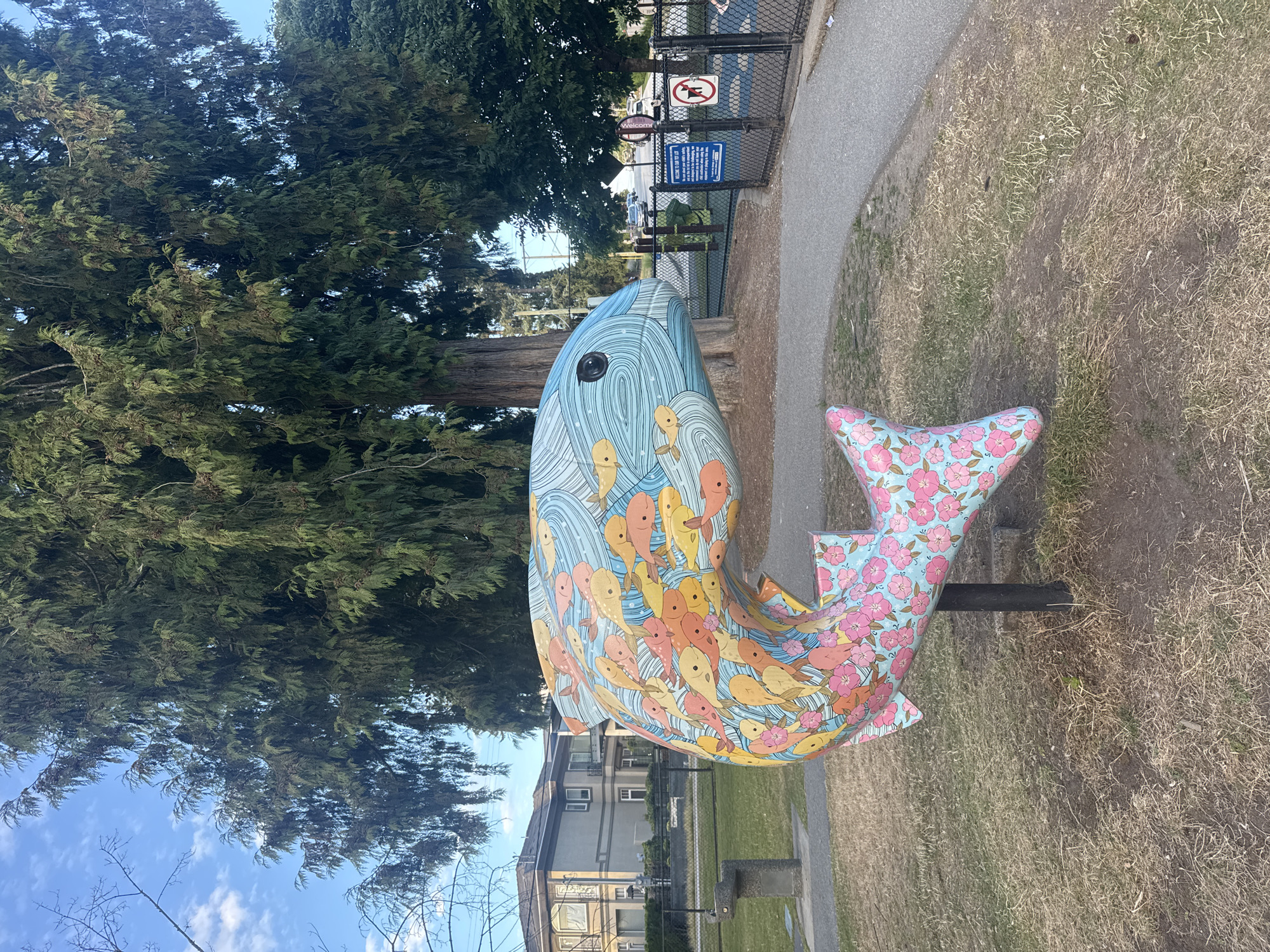 The image shows a colorful fish sculpture in a park setting. The sculpture is painted with a variety of fish in shades of orange, yellow, and blue, as well as pink flowers on a light blue background. The fish sculpture is positioned on a black pole in a grassy area. In the background, there is a large evergreen tree, a residential building, a paved path, a fence, and what appears to be a playground. A blue sign with white text and a no-dogs sign are visible on the fence. The sky is blue with scattered white clouds. The image is taken from a slightly low angle, looking up at the sculpture and the trees. The location is the City of Coquitlam, Canada.