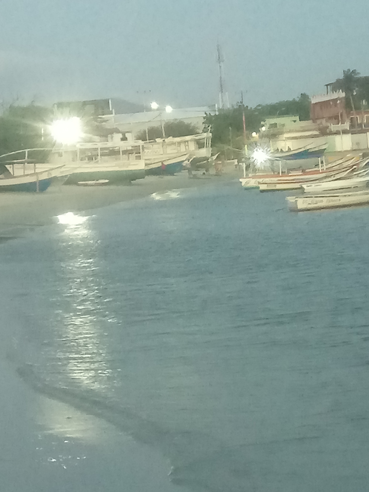 The image depicts a coastal scene at dusk in Juan Griego, Venezuela. The foreground is dominated by the ocean water reflecting light, likely from artificial sources. Several small boats are beached or moored along the shoreline in the middle ground. Buildings are visible along the shore in the background, along with a tall antenna. The overall lighting suggests either early evening or pre-dawn.