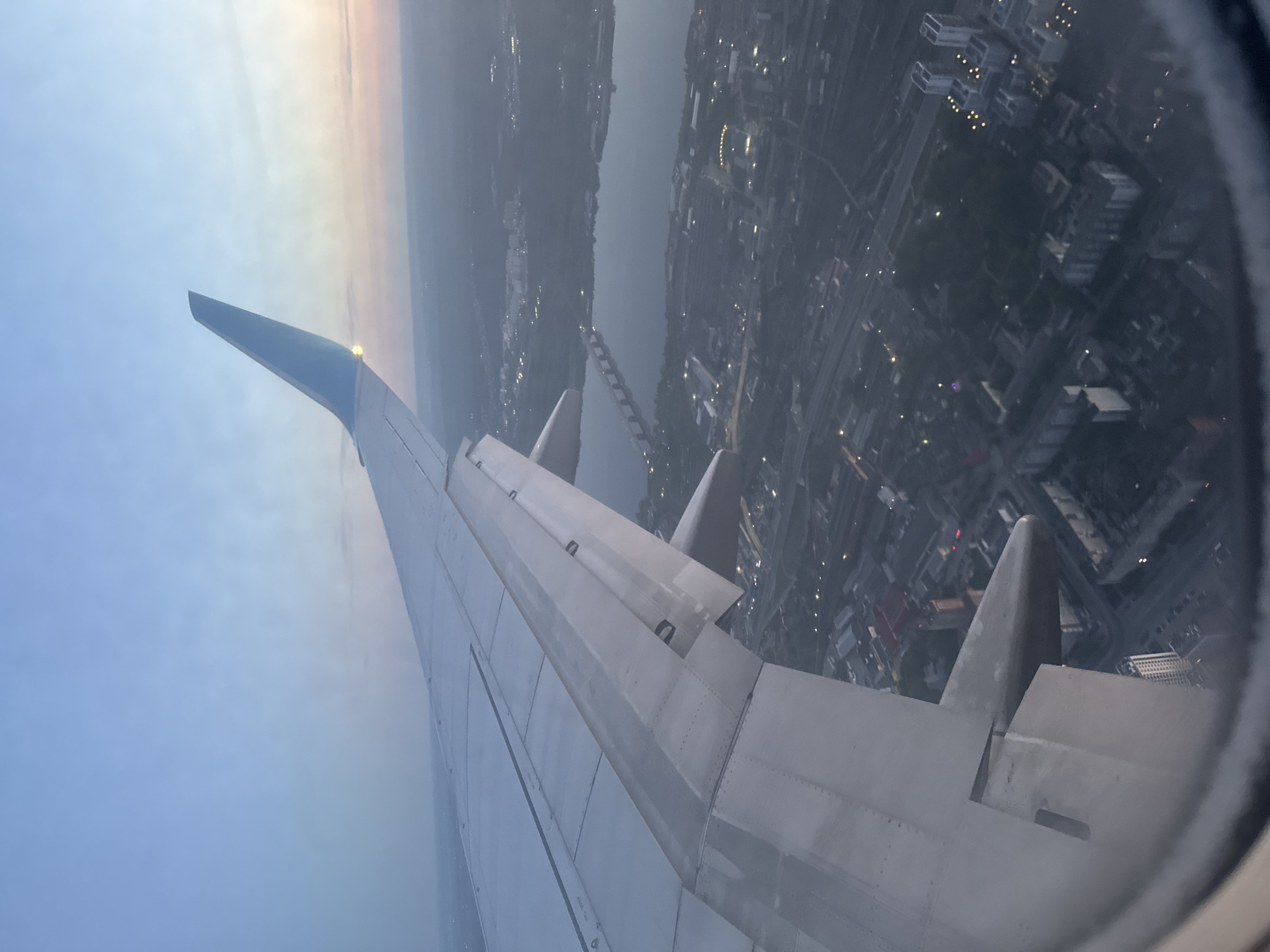 The image, taken from the window seat of an airplane, presents a view of what appears to be a city at night. The wing of the plane is prominently featured in the foreground, stretching across the majority of the frame. Beyond the wing, one can see an urban landscape illuminated by numerous lights. A river or body of water cuts through the cityscape, with a bridge visible. At the top, the sky transitions between a faint glow and what seems to be the darkness of night or early dawn. Based on the presence of a body of water, a bridge and the grid-like pattern of city lights, this could be the city of Perm, Russia, although that would need verification.