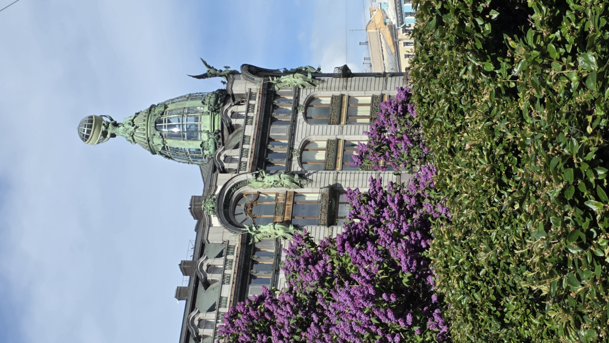 The image shows the Singer House, also known as the Dom Knigi (House of Books), in Saint Petersburg, Russia. The building features a distinctive glass dome topped with a sculpture. Part of the building's facade is visible, with windows, decorative sculptures, and purple flowers in bloom. A green hedge is visible on the right side of the frame. The sky is blue with some light clouds. The image appears to be taken from a low angle, capturing the height of the building.