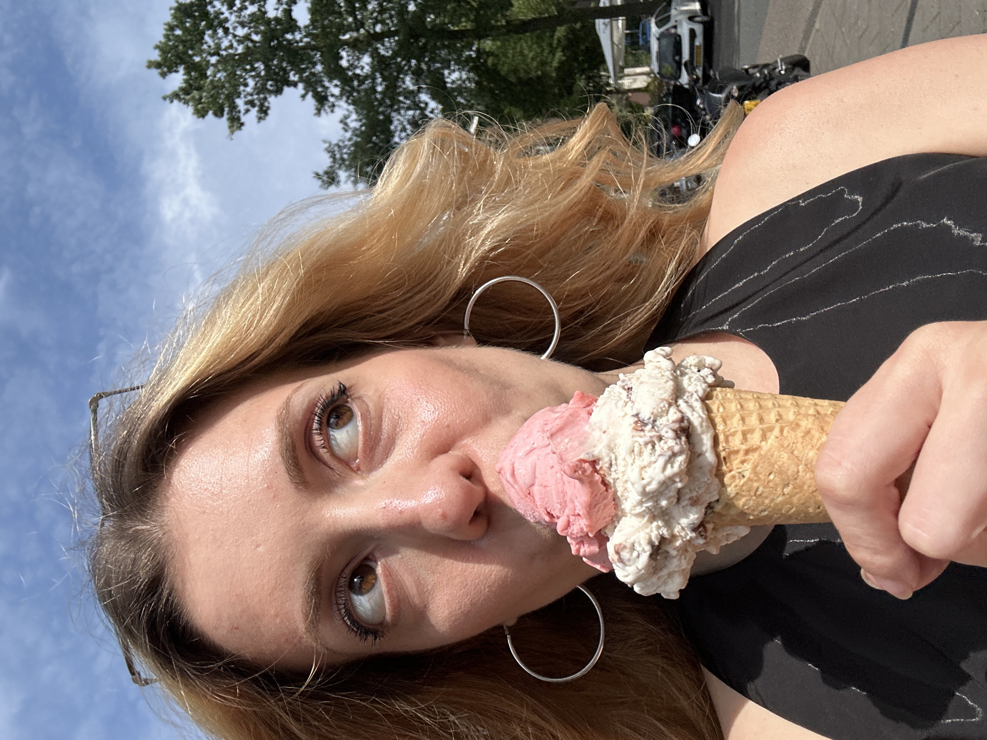 A woman with long blonde hair and silver hoop earrings is holding an ice cream cone with a scoop of pink ice cream and a scoop of white ice cream, with the pink scoop near her mouth. She has brown eyes and is wearing a black shirt. In the background, there are trees, a blue sky with clouds, and vehicles that appear to be motorcycles and a van. The location could potentially be in Amsterdam, Netherlands, but there is not enough detail in the photo to confirm that as fact.