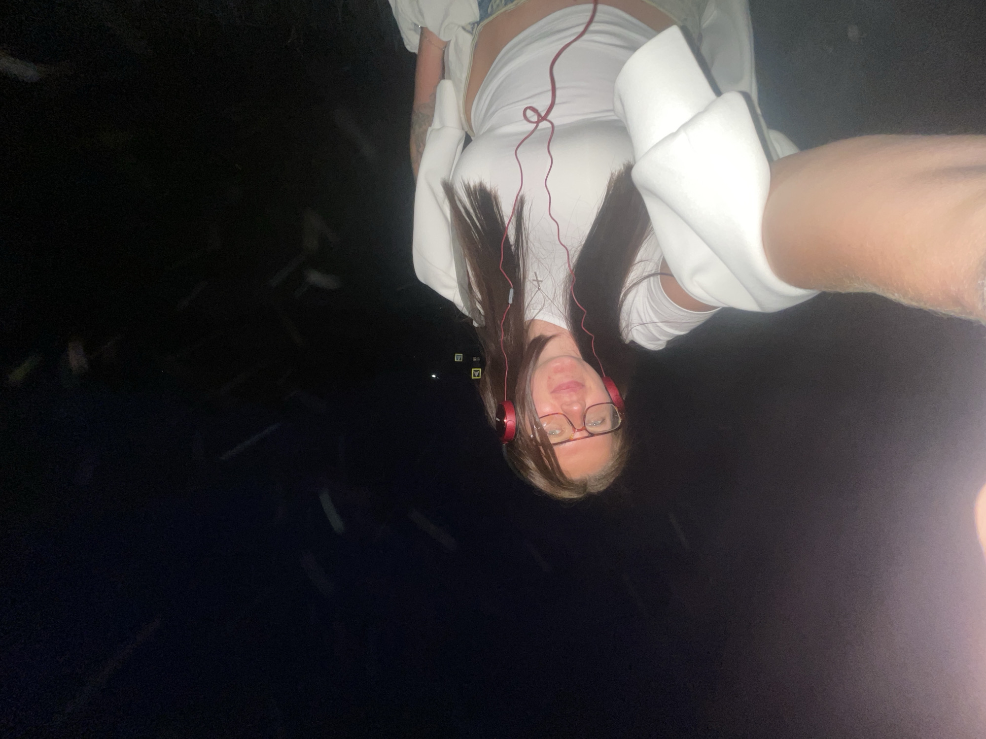The image depicts a woman with long brown hair, wearing red headphones and glasses, and a white sweatshirt. She is oriented upside down in the frame, with a dark background suggesting a night setting. The location is potentially the city of Molokovo, Russia.