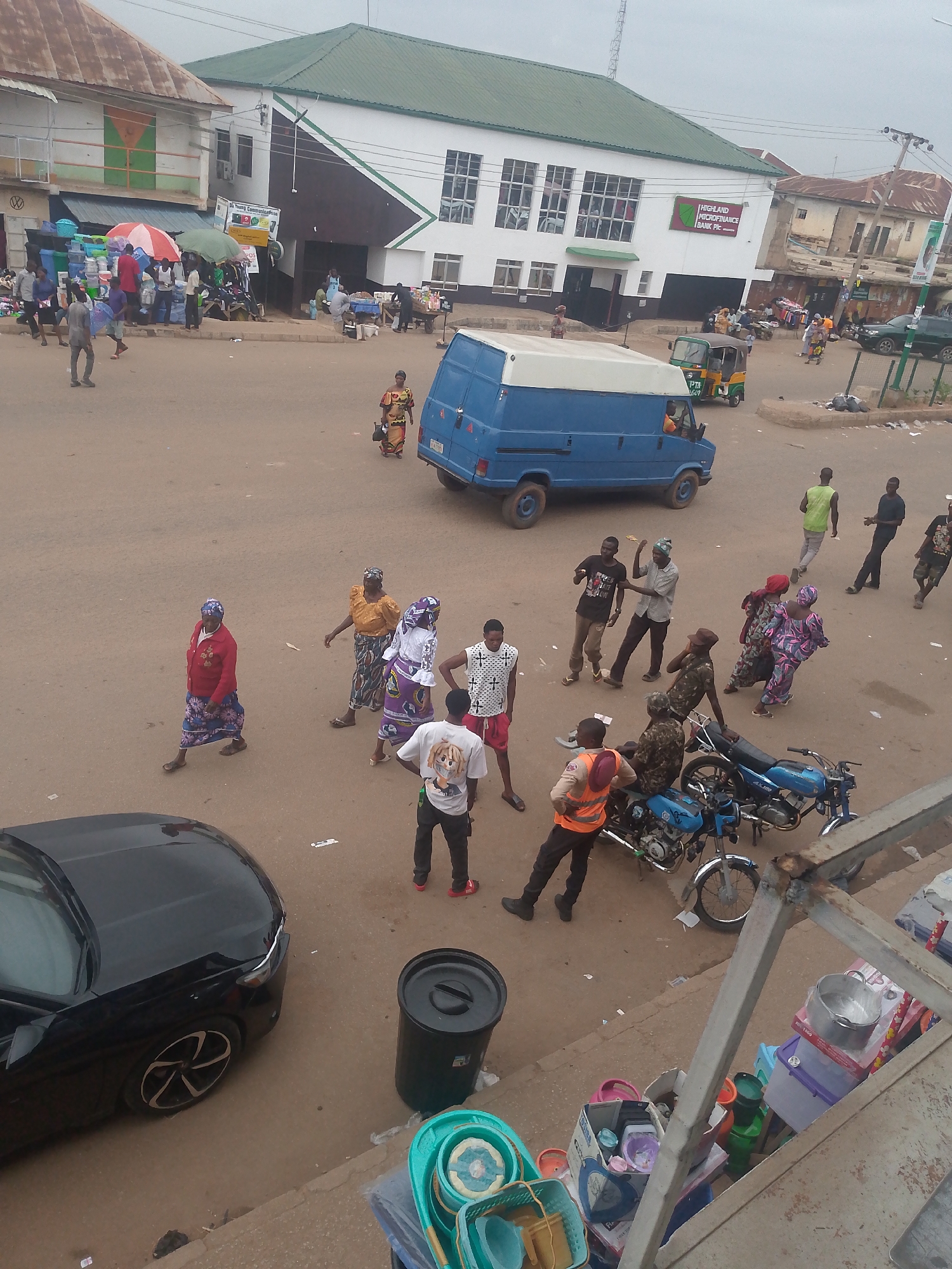 The image captures a bustling street scene in Bukuru, Nigeria, under an overcast sky.
**Environment:** The street is paved with reddish-brown soil and dust, typical of the region. Buildings flank the road, including a white building with a green roof and signage that says "Highland Microfinance Bank Plc." A smaller building with a rusty metal roof is also visible.
**People:** The street is populated with numerous individuals of African descent, of varying ages and attire. Some people are walking, while others are standing in groups. Some of the clothing details observed:
- Women in traditional colorful dresses and headscarves.
- Men in casual wear, including t-shirts and trousers.
- A man in an orange vest standing next to a motorcycle.
- Three men in military camouflage sitting on motorbikes.
**Objects:** Notable objects in the scene include:
- A blue van with a white roof parked in the middle of the street.
- Several blue motorcycles with riders.
- A black car parked along the curb.
- A black trash receptacle next to the black car
- Plastic items such as buckets and storage boxes are visible in the foreground
**Actions & Interactions:**
- People are engaged in various activities, including walking across the street, conversing in groups, and riding motorcycles.
- Some people appear to be haggling in the background.
**Overall:** The image portrays a typical day in Bukuru, Nigeria, characterized by the mix of traffic, pedestrians, and commercial activity along a busy street.