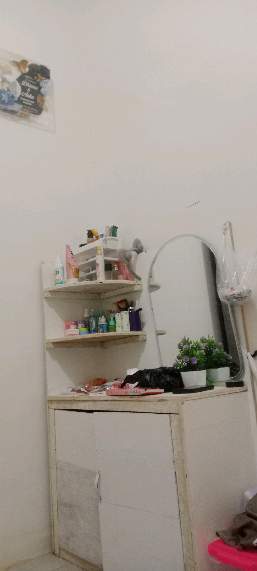 A vertical, medium shot captures a cluttered vanity area in what appears to be a bedroom or dressing room in Serongga, Indonesia. The scene is dominated by a white wall and a simple, light-colored wooden vanity cabinet with a single door. Above the cabinet, two white wooden shelves are attached to the wall, housing an assortment of toiletries and cosmetics. These include various bottles, tubes, and containers, some with colorful labels, suggesting a personal grooming space.
To the right of the shelves, a large, oval-shaped mirror reflects a portion of the room, though details are indistinct due to the angle and lighting. On the countertop of the vanity, a black plastic bag is prominent, along with two small potted plants with purple flowers. A few other items, including what looks like a pink box or book, are also visible on the vanity surface.
On the far left of the frame, a small, transparent plastic package with a floral design and text that is partially legible, potentially a wedding favor or gift, is affixed to the wall. The overall impression is one of a functional, albeit somewhat disorganized, personal space, typical of a domestic setting in Serongga. The lighting is diffuse, creating soft shadows, and the color palette is muted, with whites and light wood tones prevailing.