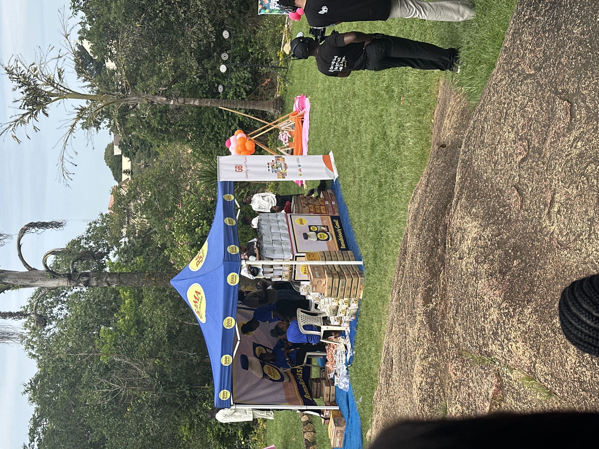 The image depicts an outdoor event, possibly a product launch or promotional activity, set in a green, grassy area with large rocks in the foreground. In the mid-ground, a blue canopy tent with yellow branding is set up, displaying what appears to be food or beverage products. Several people are visible under and around the tent, some interacting with the products or each other, while others stand nearby. Behind the tent, a tall, thin tree with drooping fronds stands out against the background foliage and buildings. To the right of the tent, there is a small setup with balloons, a wooden frame, and a pink blanket, suggesting a picnic or relaxation area. Further to the right, two individuals wearing black t-shirts with some writing and one person in light-colored pants are partially visible, one of them appearing to be operating a video camera. The environment suggests a warm climate, with lush greenery and tropical-looking trees. The presence of promotional tents, product displays, and media equipment indicates a public gathering or event. The rocks in the foreground are large and have a reddish-brown, textured surface, with some small patches of grass growing in crevices. The sky is overcast, with a hazy appearance. This scene could plausibly be located in Jos, Nigeria, given the vegetation and architectural hints in the background, suggesting a natural, park-like setting common in some urban or peri-urban areas of the city.