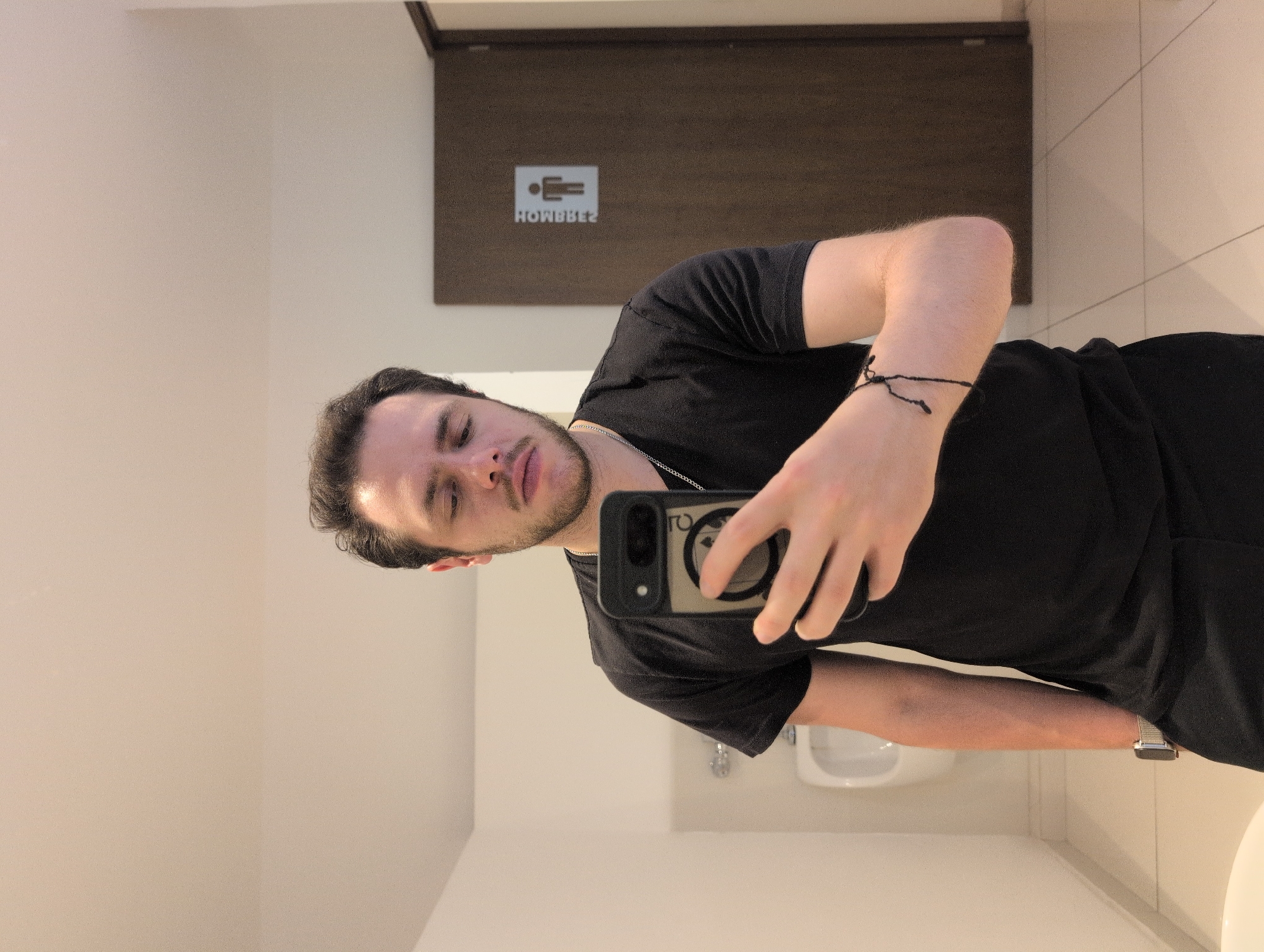A fair-skinned man is taking a selfie in a restroom with a sign for “Hombres” above his head. He is wearing a black t-shirt, a silver necklace and a thin black bracelet on his left wrist. He is holding a smartphone in his right hand. The restroom has a white sink and white tile on the walls. The photograph was taken at an unknown location in Jesús del Monte, Mexico.