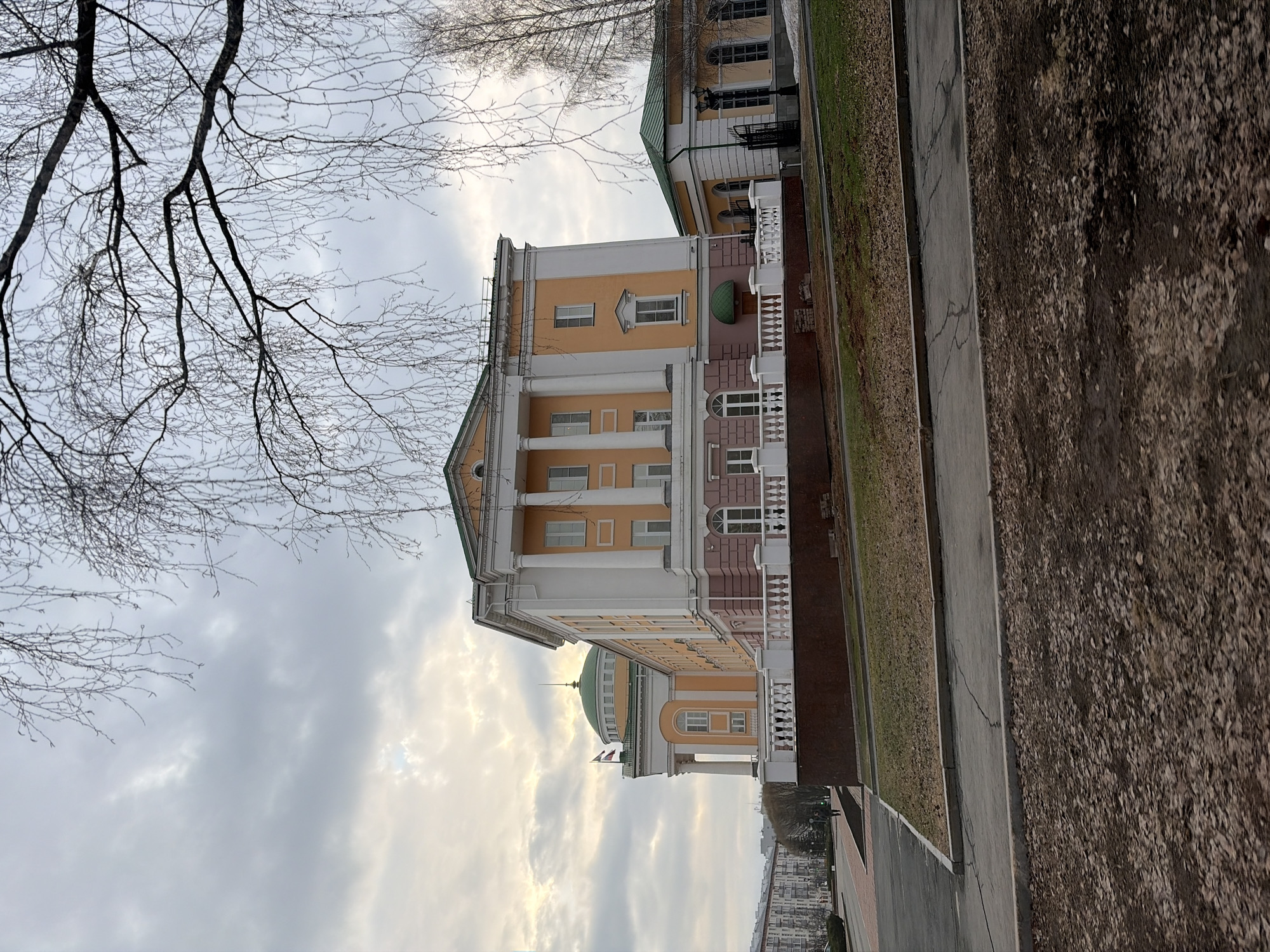 The image depicts a building with classical architecture, featuring columns, a dome, and light yellow and white facades. It appears to be situated in Izhevsk, Russia. A bare tree branch is visible in the upper left, against a cloudy sky. A sidewalk and adjacent landscaping are in the foreground, sloping slightly upward to the right. In the distance, multiple buildings are visible.