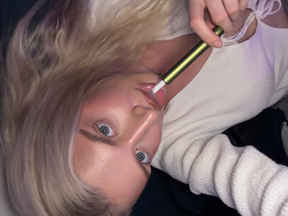 A close-up, angled portrait features a young adult female with blonde hair and blue eyes, looking directly at the camera. She is positioned as if reclining or leaning, with her head tilted. Her left hand holds an olive-green, slender, cylindrical device with a white tip, placed against her slightly parted, glossed lips, indicating active use of a vaping or heated tobacco product. She wears a light-colored, textured, long-sleeved top, possibly white or off-white, with a tie-front detail visible on the chest. Her face exhibits distinct makeup, including prominent blush on her cheeks. The background is dark and out of focus, offering no discernible environmental details. The specific location within Belgrade, Serbia, cannot be determined from the image.