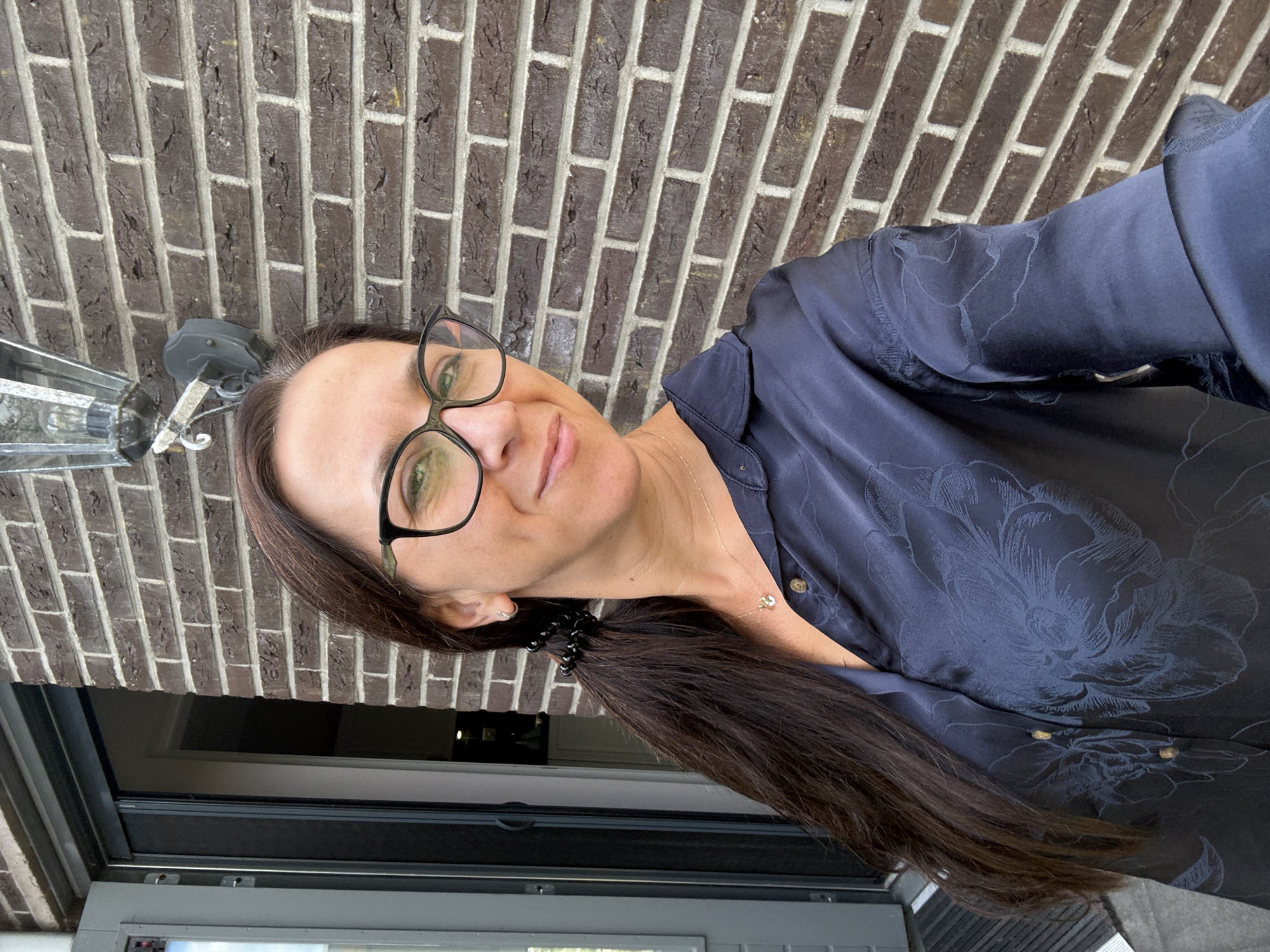 A woman with long, dark hair pulled into a ponytail, wearing glasses and a dark blue collared shirt with a floral pattern, stands in front of a brick wall. She is smiling and looking towards the camera. To her left, a glass-paned door is visible, partially open, revealing an interior space. An outdoor light fixture is mounted on the brick wall above and to her right. The scene appears to be an exterior residential setting.