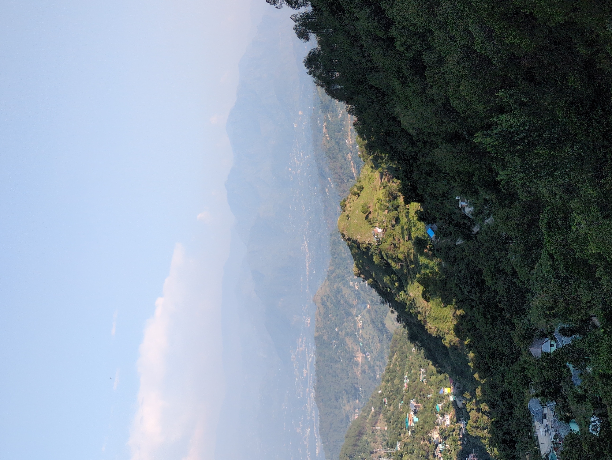 This is a wide, high-angle shot of a mountainous landscape with a town nestled within it. The sky is a pale blue with scattered white clouds. In the foreground, a steep, tree-covered hillside dominates the right side of the frame. The trees are a dark green and appear to be a mix of pine and other deciduous trees. Sunlight casts shadows across the hillside.
In the mid-ground, a smaller, greener hill slopes downwards to the left. There are scattered buildings with red and blue roofs visible on this hill, suggesting a small settlement. A few tall communication towers are also present. Further in the distance, layers of hazy mountains recede into the background, with what appears to be a larger town or city spread across the valleys. The buildings in this distant area are small and numerous, giving a sense of urban sprawl within the mountainous terrain. The overall impression is a rural or semi-rural environment in a hilly region, likely in or around Doh, India, given the context. There are no visible people or specific actions taking place. The environment is natural with man-made structures integrated into the landscape.