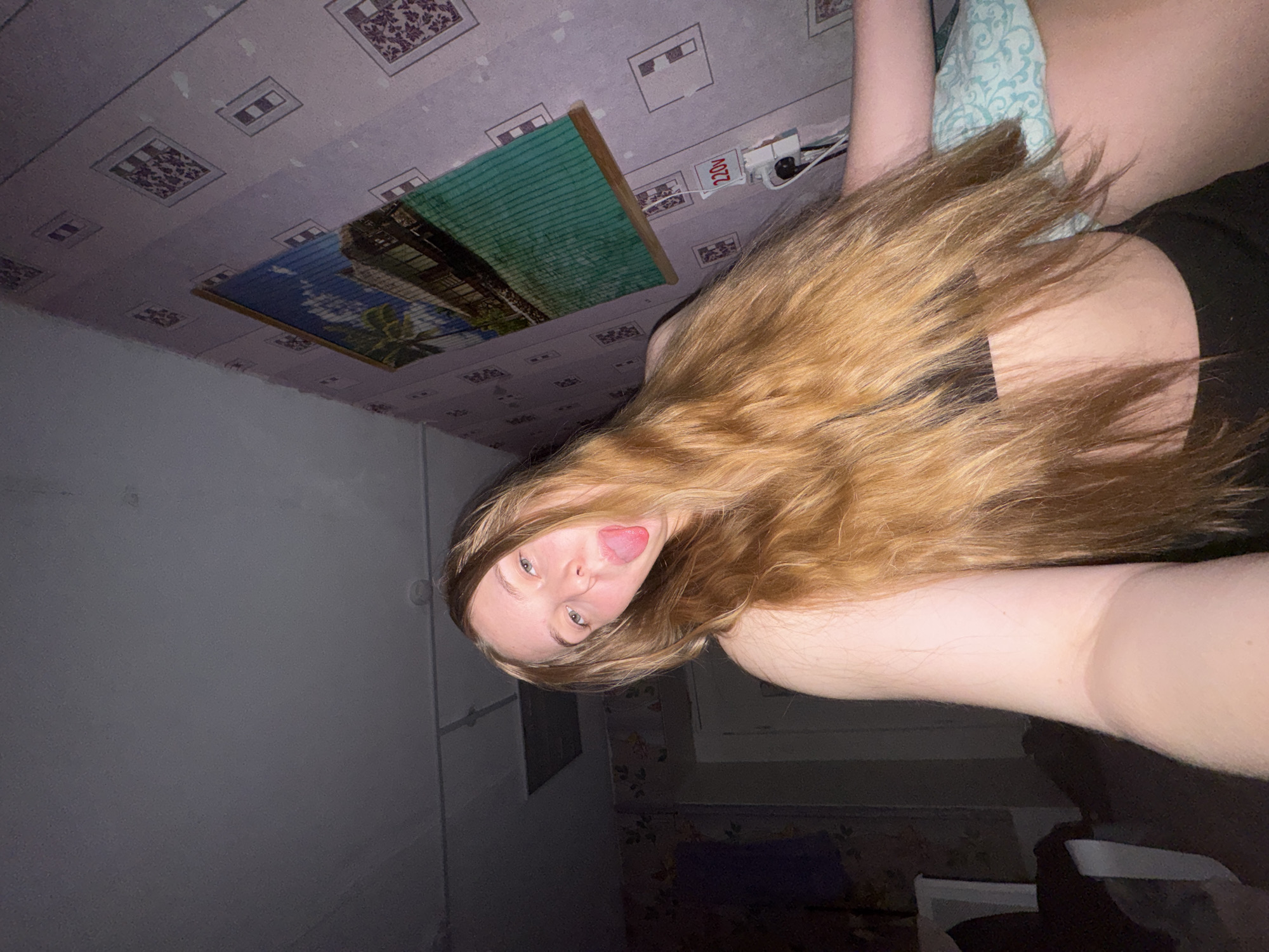 The image depicts a young Caucasian female in an indoor residential setting, identified as Perm, Russia. The female, with long, light brown hair and light-colored eyes, is shown from the chest up, extending her tongue towards the camera. She is wearing a dark brassiere or sports top and dark-colored bottoms. The immediate background on the right features a wall covered with light purple wallpaper displaying repeating square patterns, which appear worn in areas. A framed picture depicting a tropical scene with palm trees and a building is mounted on this wall. Below the picture, a white electrical outlet with a visible plug is present, and the voltage "220V" is marked on the wall or the outlet itself. The ceiling is a uniform grey. To the left, a darker wall with a faded floral wallpaper pattern is visible, along with a partial door frame. A light blue patterned blanket or pillow is partially visible on the lower right, beneath the subject's arm. The lighting is dim, suggesting an internal light source, possibly a phone flash, illuminating the subject directly.
